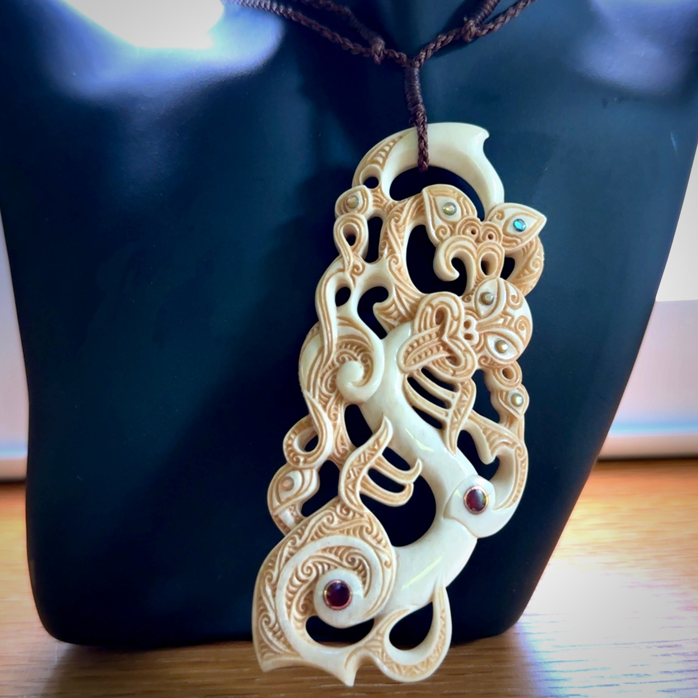 This picture shows a carved Tiki and manaia in bone. The artist, Andrew Doughty, has carved traditional decorative Koru designs into the face of the body and these run up the sides of the manaia head. These have specific meanings. It is provided with a hand-plaited brown cord that is length adjustable.