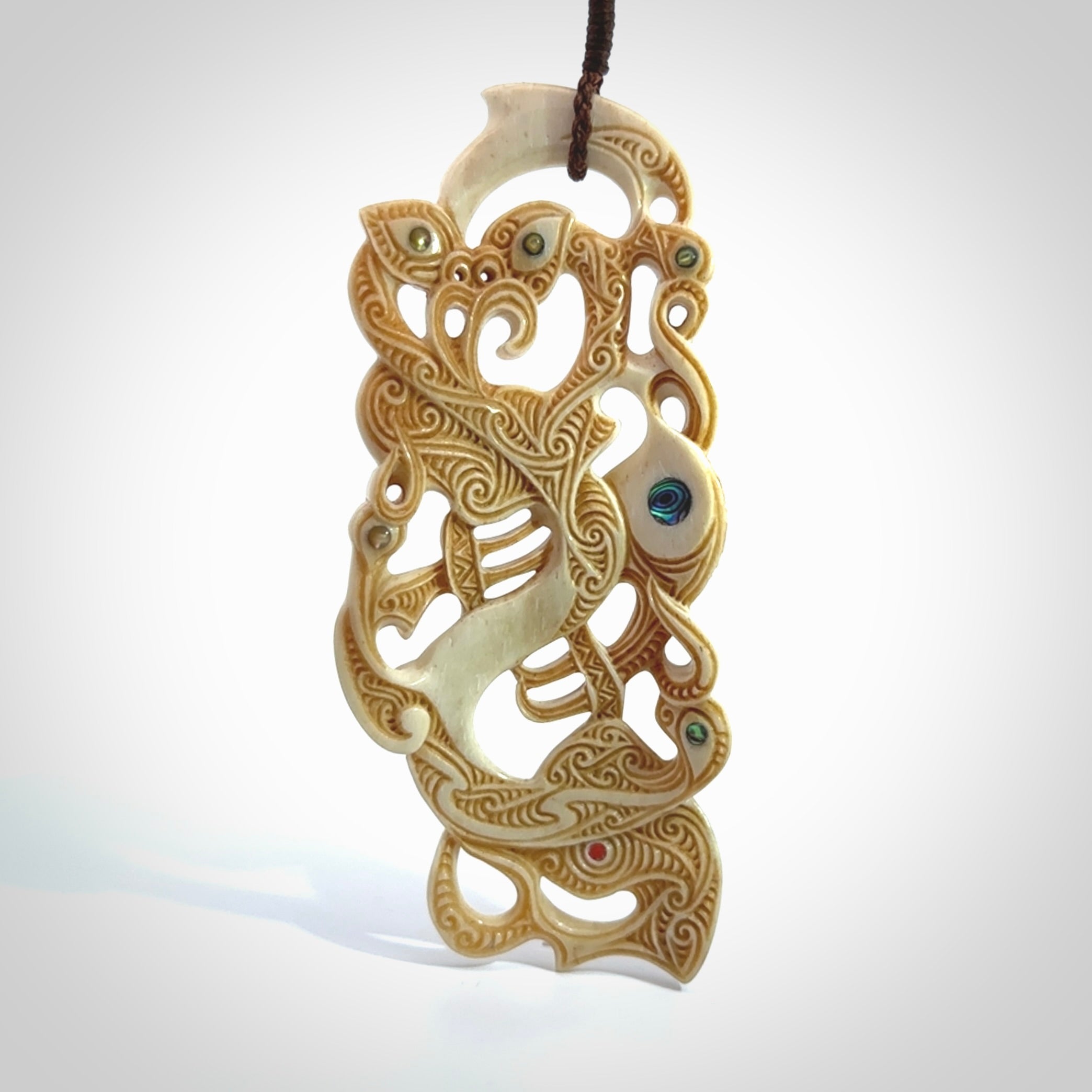 This picture shows a carved Tiki and manaia in bone. The artist, Andrew Doughty, has carved traditional decorative Koru designs into the face of the body and these run up the sides of the manaia head. These have specific meanings. It is provided with a hand-plaited brown cord that is length adjustable.