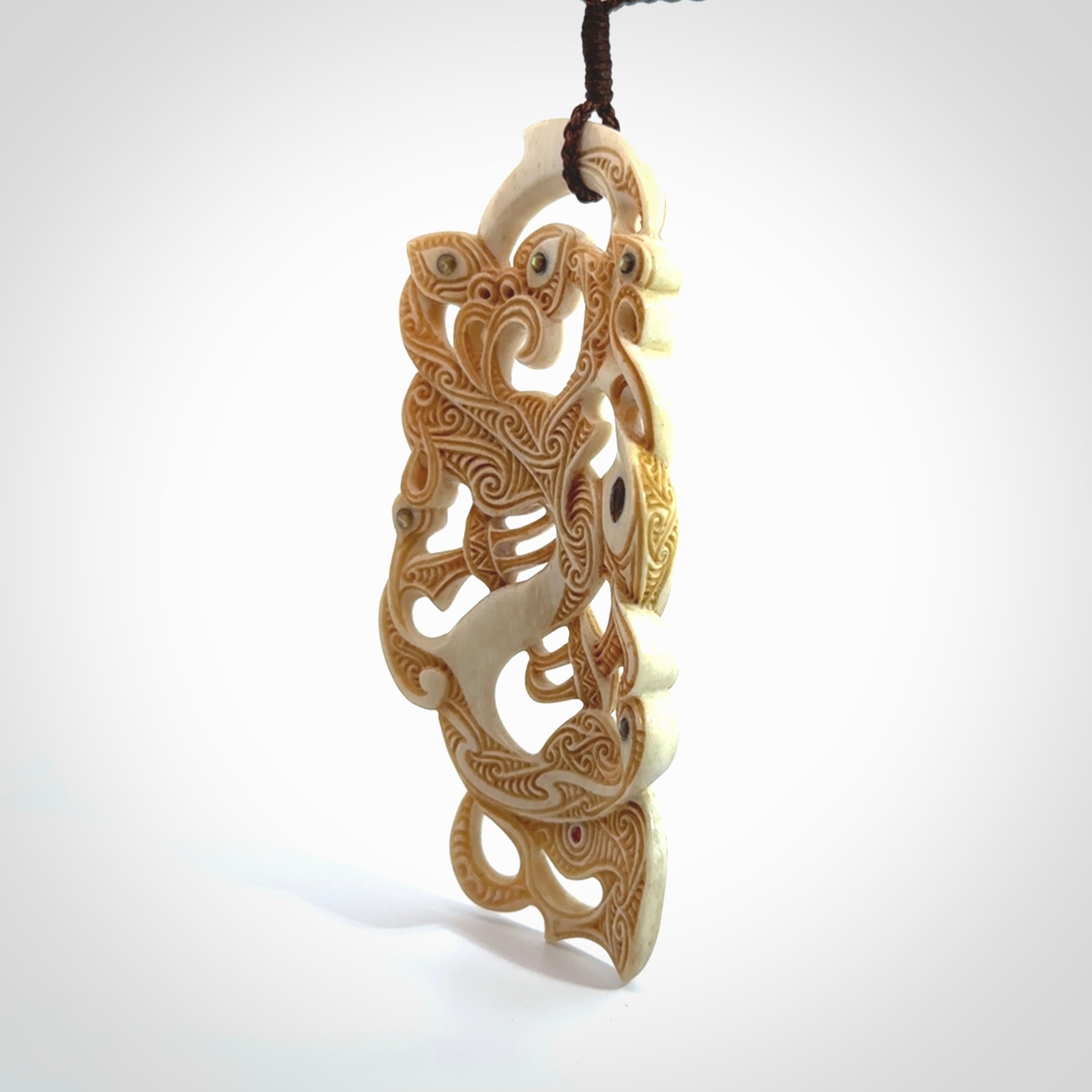 This picture shows a carved Tiki and manaia in bone. The artist, Andrew Doughty, has carved traditional decorative Koru designs into the face of the body and these run up the sides of the manaia head. These have specific meanings. It is provided with a hand-plaited brown cord that is length adjustable.