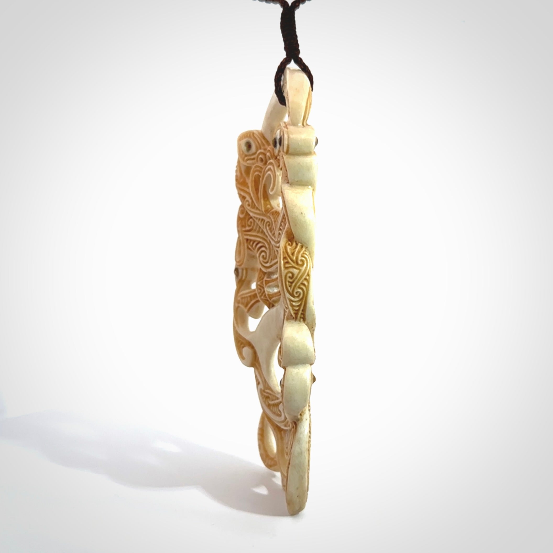 This picture shows a carved Tiki and manaia in bone. The artist, Andrew Doughty, has carved traditional decorative Koru designs into the face of the body and these run up the sides of the manaia head. These have specific meanings. It is provided with a hand-plaited brown cord that is length adjustable.