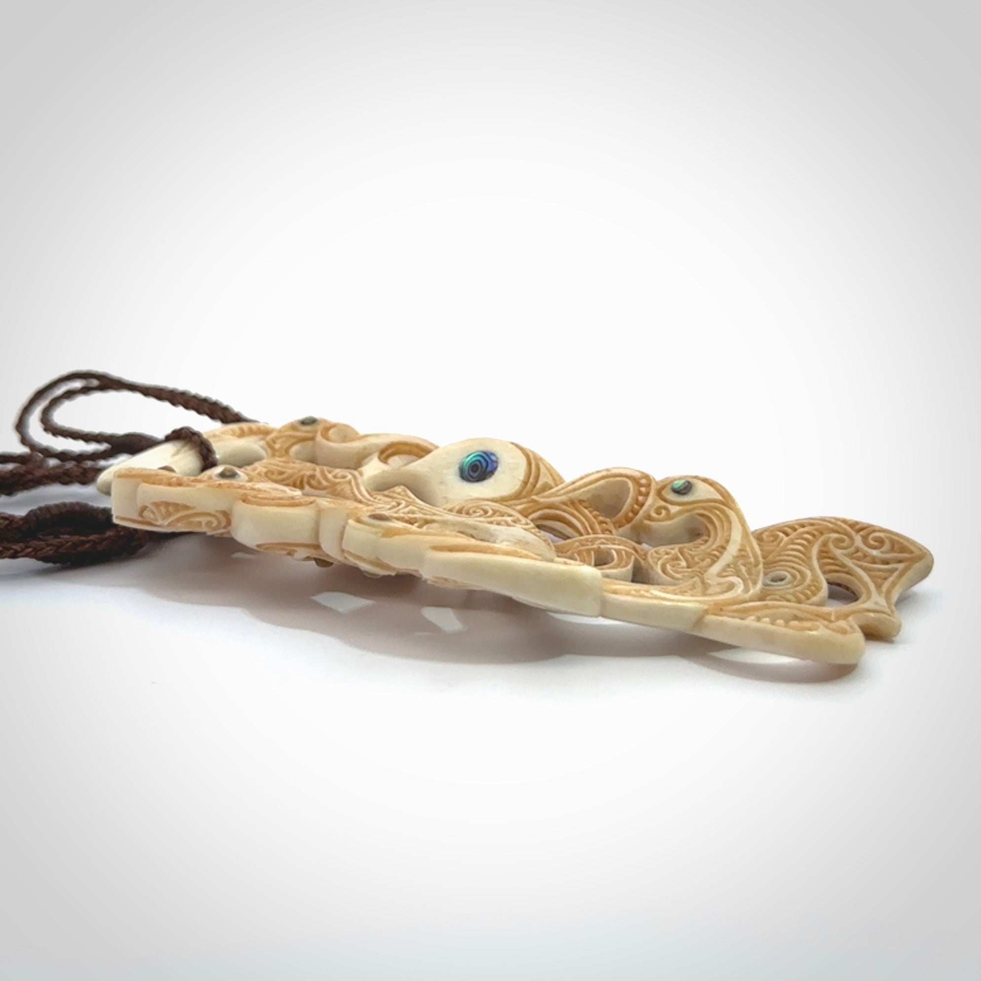 This picture shows a carved Tiki and manaia in bone. The artist, Andrew Doughty, has carved traditional decorative Koru designs into the face of the body and these run up the sides of the manaia head. These have specific meanings. It is provided with a hand-plaited brown cord that is length adjustable.