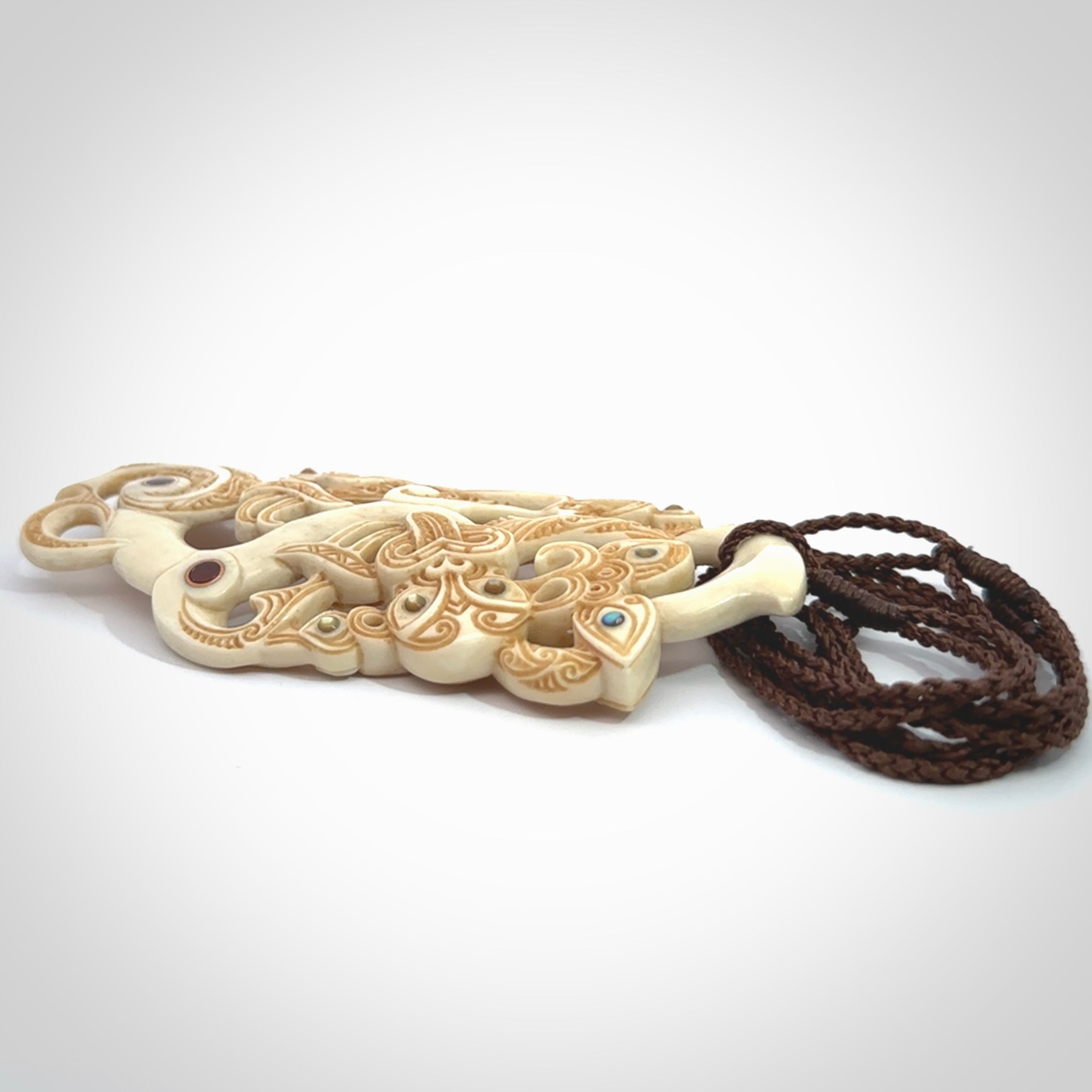 This picture shows a carved Tiki and manaia in bone. The artist, Andrew Doughty, has carved traditional decorative Koru designs into the face of the body and these run up the sides of the manaia head. These have specific meanings. It is provided with a hand-plaited brown cord that is length adjustable.