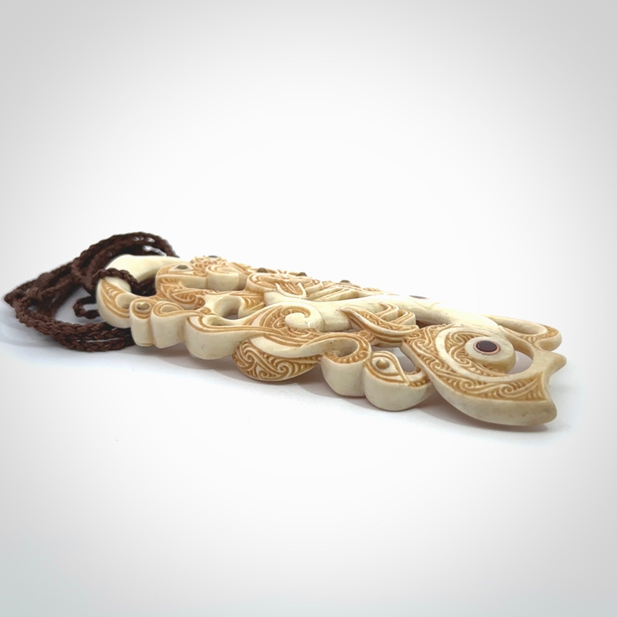 This picture shows a carved Tiki and manaia in bone. The artist, Andrew Doughty, has carved traditional decorative Koru designs into the face of the body and these run up the sides of the manaia head. These have specific meanings. It is provided with a hand-plaited brown cord that is length adjustable.