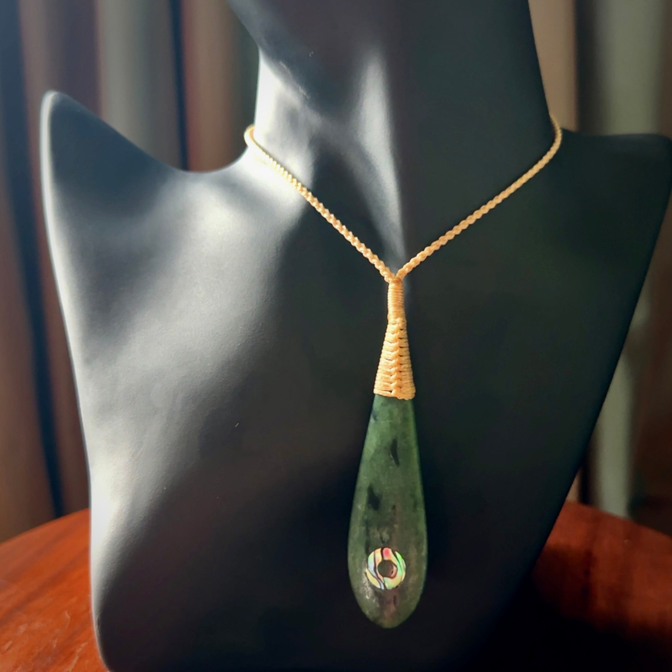 This photo shows a beautiful jade drop with paua shell inlay. It is hand carved from a piece of green New Zealand pounamu, with fascinating light green inclusions throughout the stone. We have this on a beige coloured, adjustable four plait cord. We ship this worldwide with an express courier service. Postage is free.