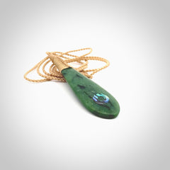 This photo shows a beautiful jade drop with paua shell inlay. It is hand carved from a piece of green New Zealand pounamu, with fascinating light green inclusions throughout the stone. We have this on a beige coloured, adjustable four plait cord. We ship this worldwide with an express courier service. Postage is free.