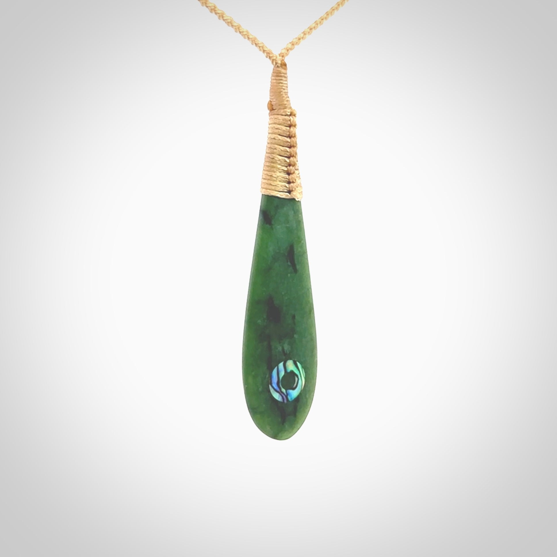 This photo shows a beautiful jade drop with paua shell inlay. It is hand carved from a piece of green New Zealand pounamu, with fascinating light green inclusions throughout the stone. We have this on a beige coloured, adjustable four plait cord. We ship this worldwide with an express courier service. Postage is free.