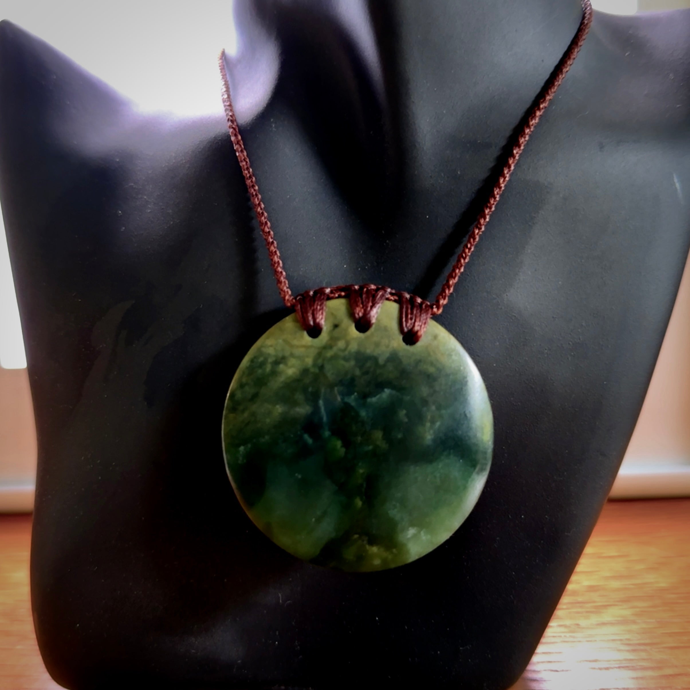 This piece is a medium sized, oval round, disc pendant. It was carved for us by Ric Moor from a lovely deep green and orange piece of New Zealand flower jade. It is suspended on an brown coloured braided cord that is length adjustable.