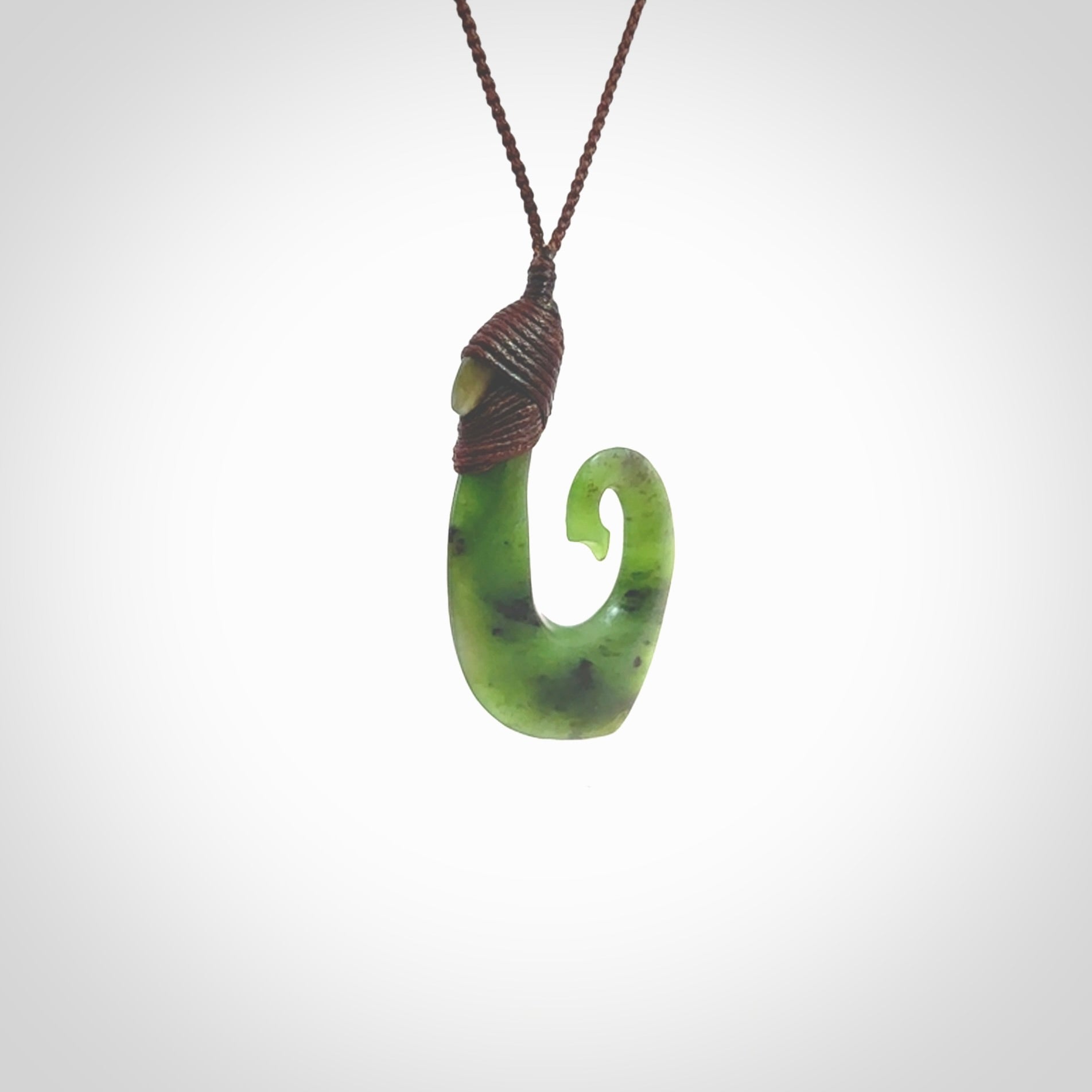 This picture shows a jade hook pendant, also called a hei-matau, carved for us in New Zealand jade. The jade is a wonderful green pounamu. This is a rare jade loved and valued for its distinctive colour. The carver is Ric Moor - and this is a beautiful example of his work. The cord is a four-plait, adjustable brown coloured necklace.