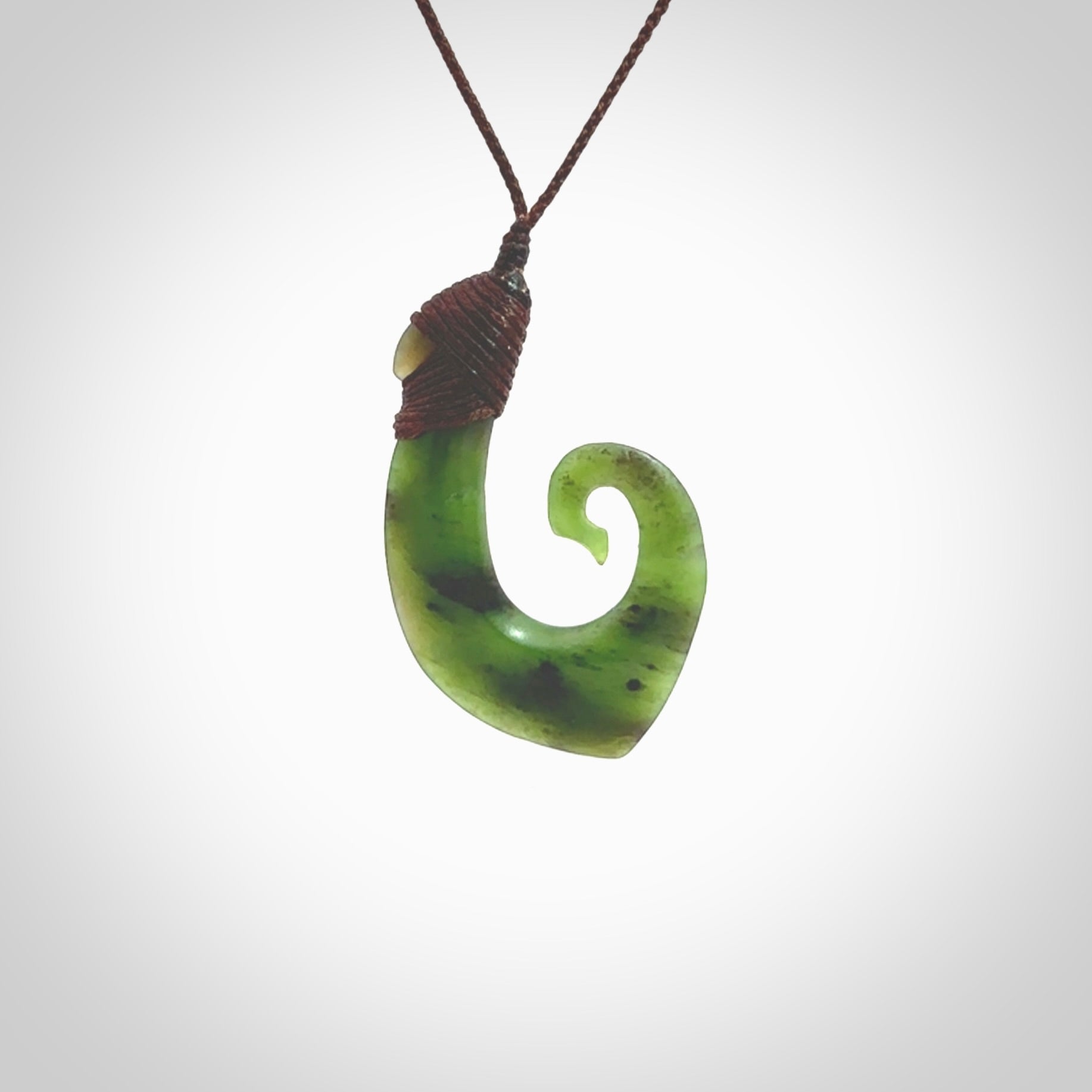 This picture shows a jade hook pendant, also called a hei-matau, carved for us in New Zealand jade. The jade is a wonderful green pounamu. This is a rare jade loved and valued for its distinctive colour. The carver is Ric Moor - and this is a beautiful example of his work. The cord is a four-plait, adjustable brown coloured necklace.