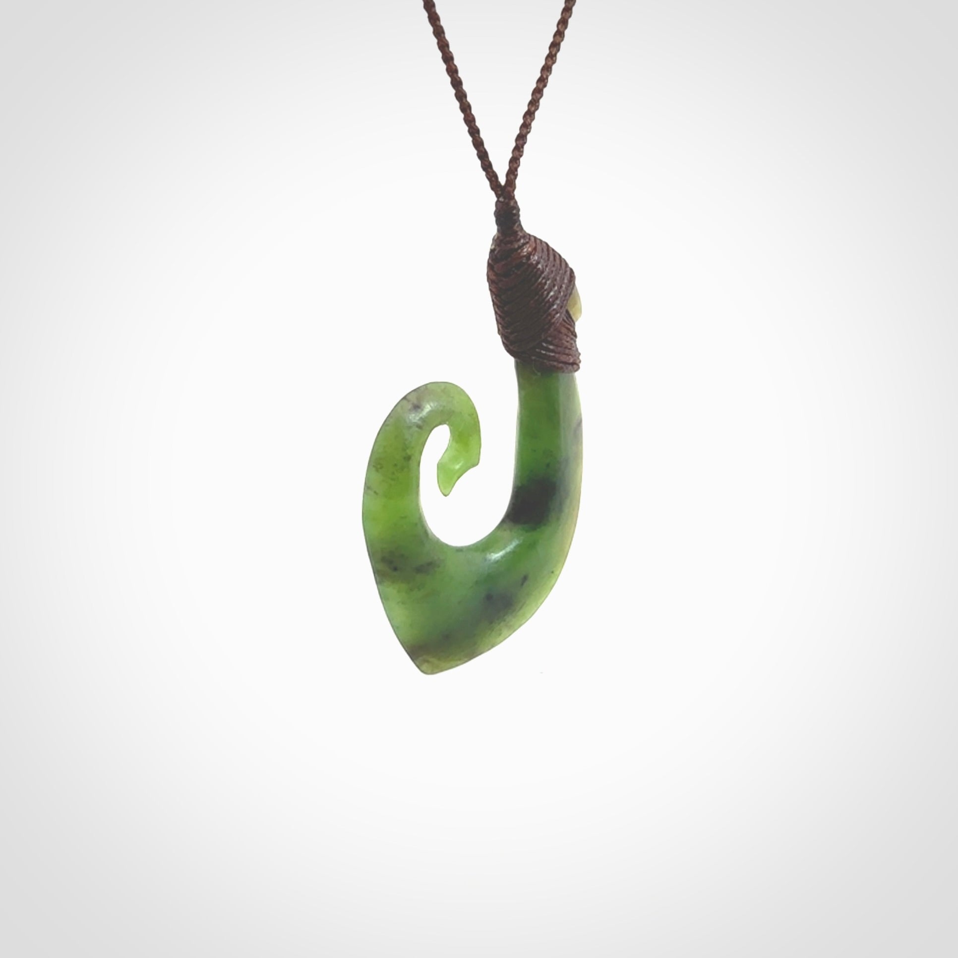 This picture shows a jade hook pendant, also called a hei-matau, carved for us in New Zealand jade. The jade is a wonderful green pounamu. This is a rare jade loved and valued for its distinctive colour. The carver is Ric Moor - and this is a beautiful example of his work. The cord is a four-plait, adjustable brown coloured necklace.