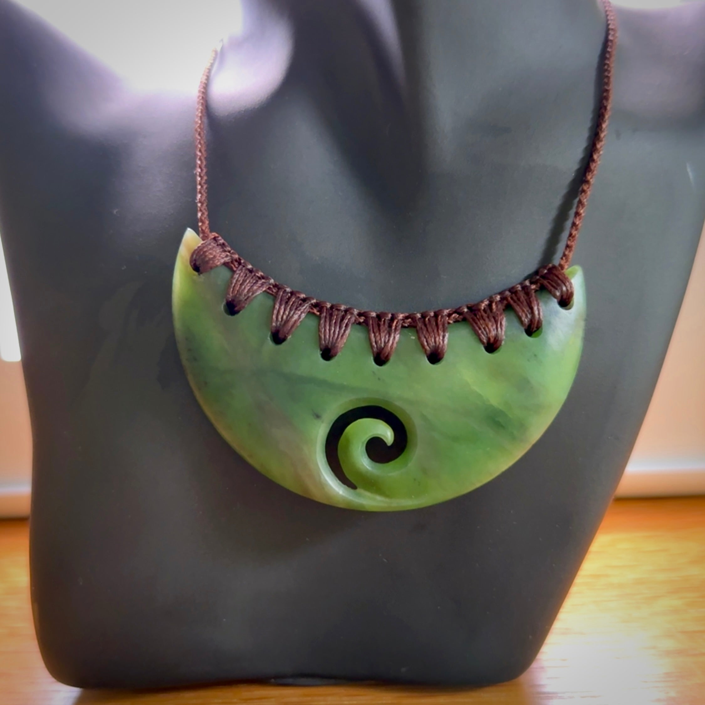 This picture shows a hand carved jade half-shield with Koru pendant. It is a semi-translucent green colour. This is a wonderful piece of jewellery. The cord is hand plaited and adjustable so that you can position the pendant where it suits you best. This piece was carved for us by Ric Moor. Delivery is free worldwide.