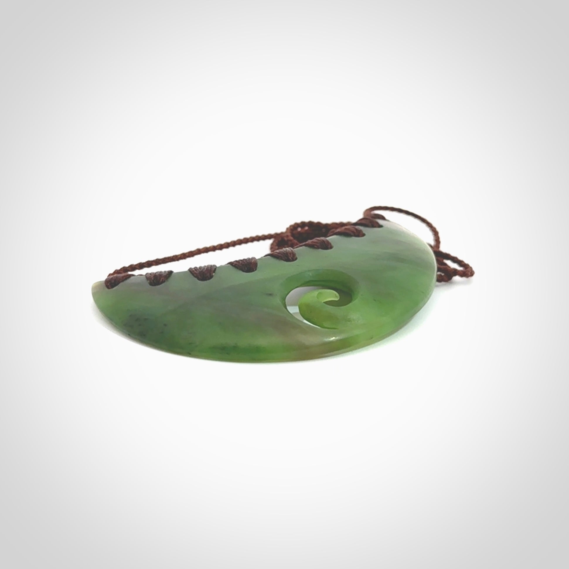 This picture shows a hand carved jade half-shield with Koru pendant. It is a semi-translucent green colour. This is a wonderful piece of jewellery. The cord is hand plaited and adjustable so that you can position the pendant where it suits you best. This piece was carved for us by Ric Moor. Delivery is free worldwide.