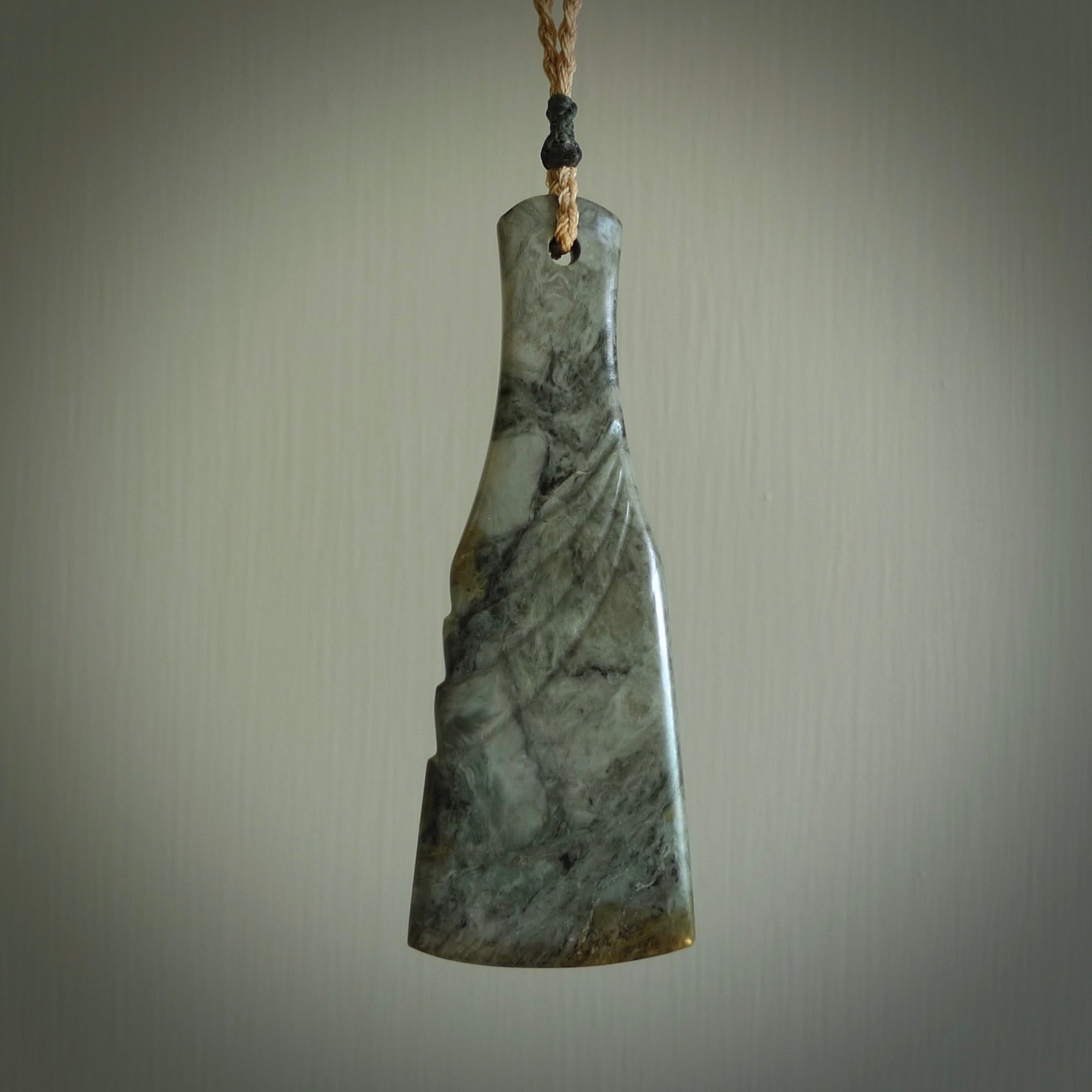 This is a sleek and slender hand carved New Zealand Jade mere pendant. The stone is a very dark green with mottled pale inclusions. The cord is tan and adjustable. The pendant has a light polish and just glows.