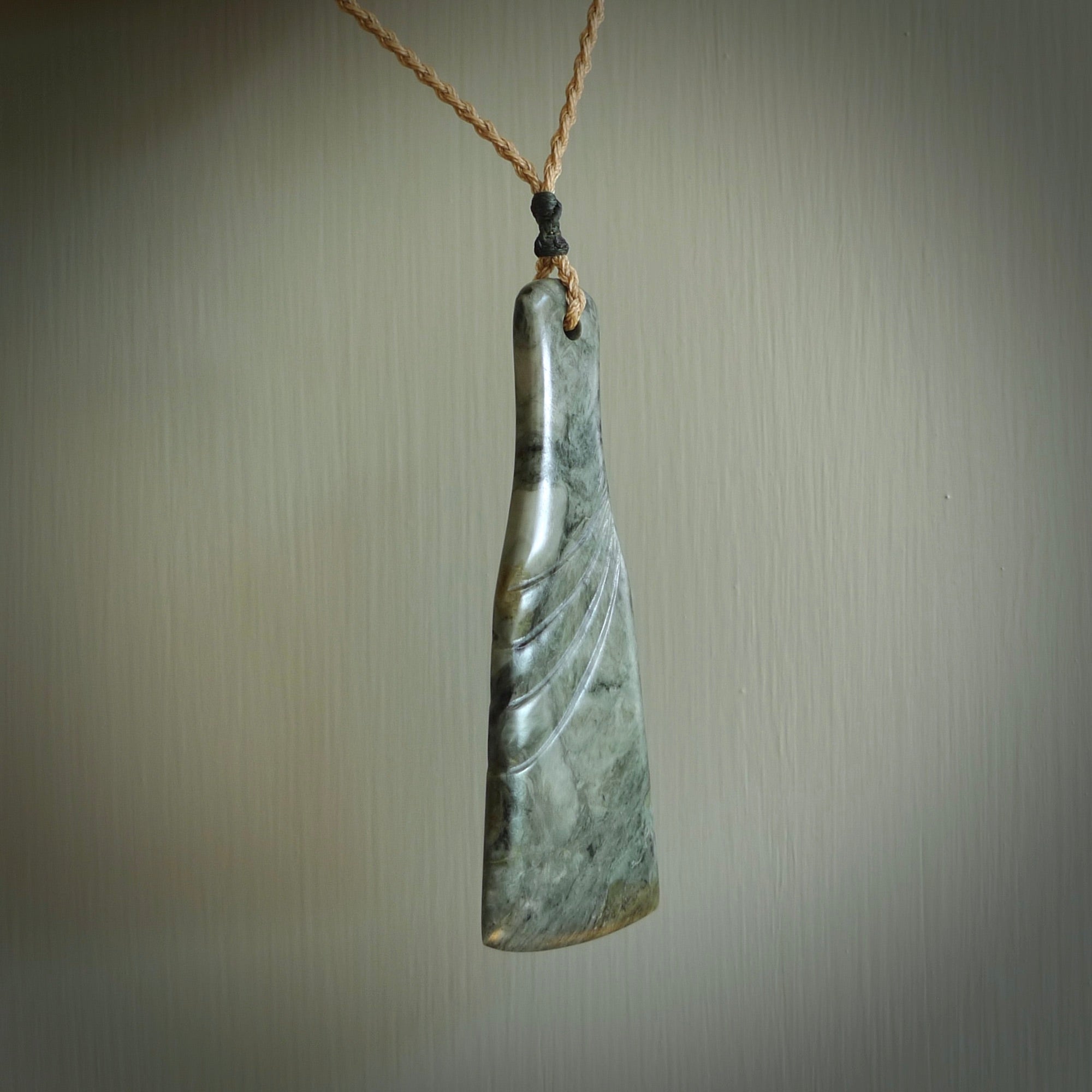 This is a sleek and slender hand carved New Zealand Jade mere pendant. The stone is a very dark green with mottled pale inclusions. The cord is tan and adjustable. The pendant has a light polish and just glows.