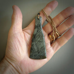 This is a sleek and slender hand carved New Zealand Jade mere pendant. The stone is a very dark green with mottled pale inclusions. The cord is tan and adjustable. The pendant has a light polish and just glows.