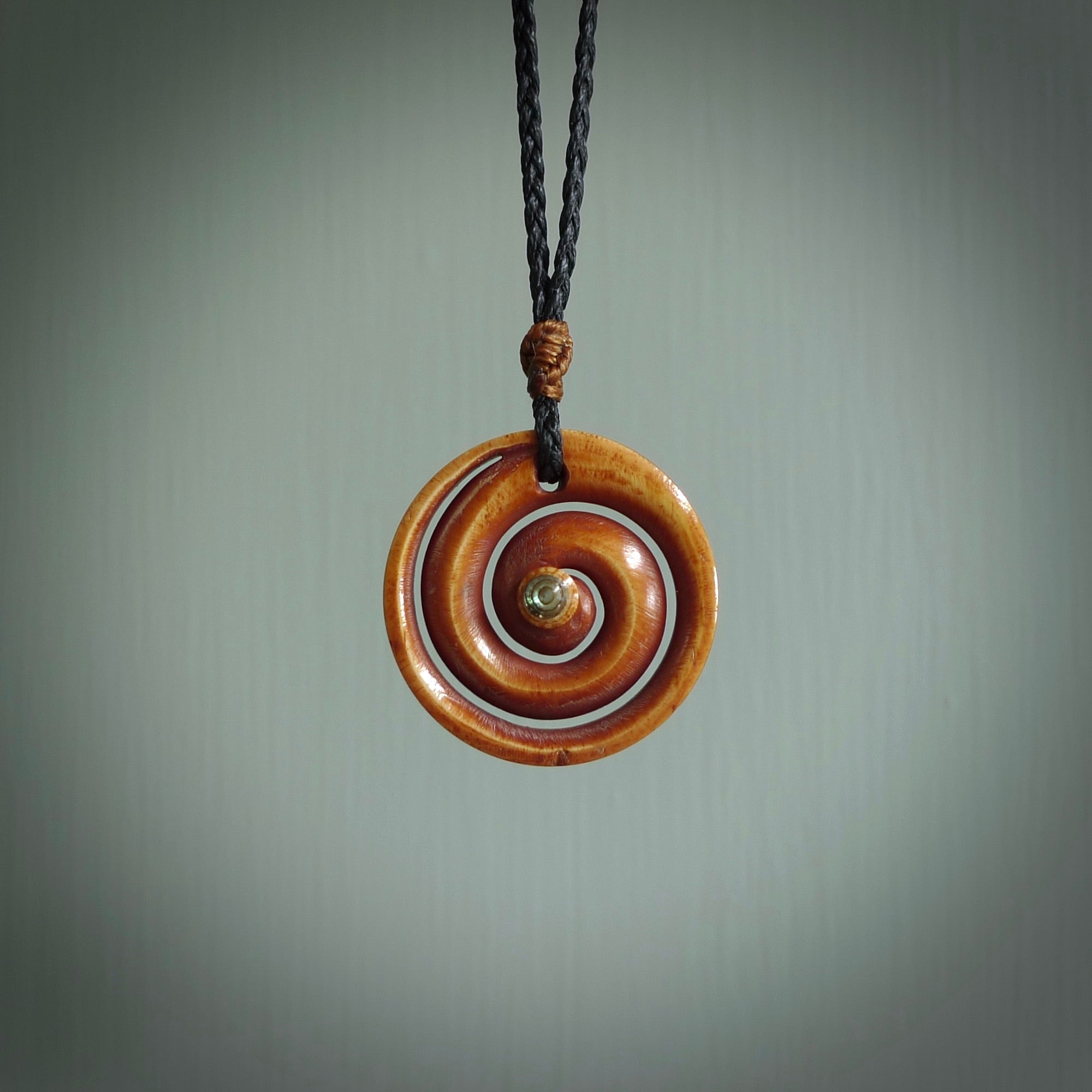 A hand carved and intricate koru pendant made for us by Yuri Terenyi. This is a beautiful little piece and is emblematic of the well known and loved Koru design. It is carved from stained bone in a hollowed, koru spiral shape with paua shell inlay in the middle of the koru. It is suspended from a black cord with a gingernut floret and the necklace is adjustable.