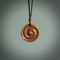 A hand carved and intricate koru pendant made for us by Yuri Terenyi. This is a beautiful little piece and is emblematic of the well known and loved Koru design. It is carved from stained bone in a hollowed, koru spiral shape with paua shell inlay in the middle of the koru. It is suspended from a black cord with a gingernut floret and the necklace is adjustable.