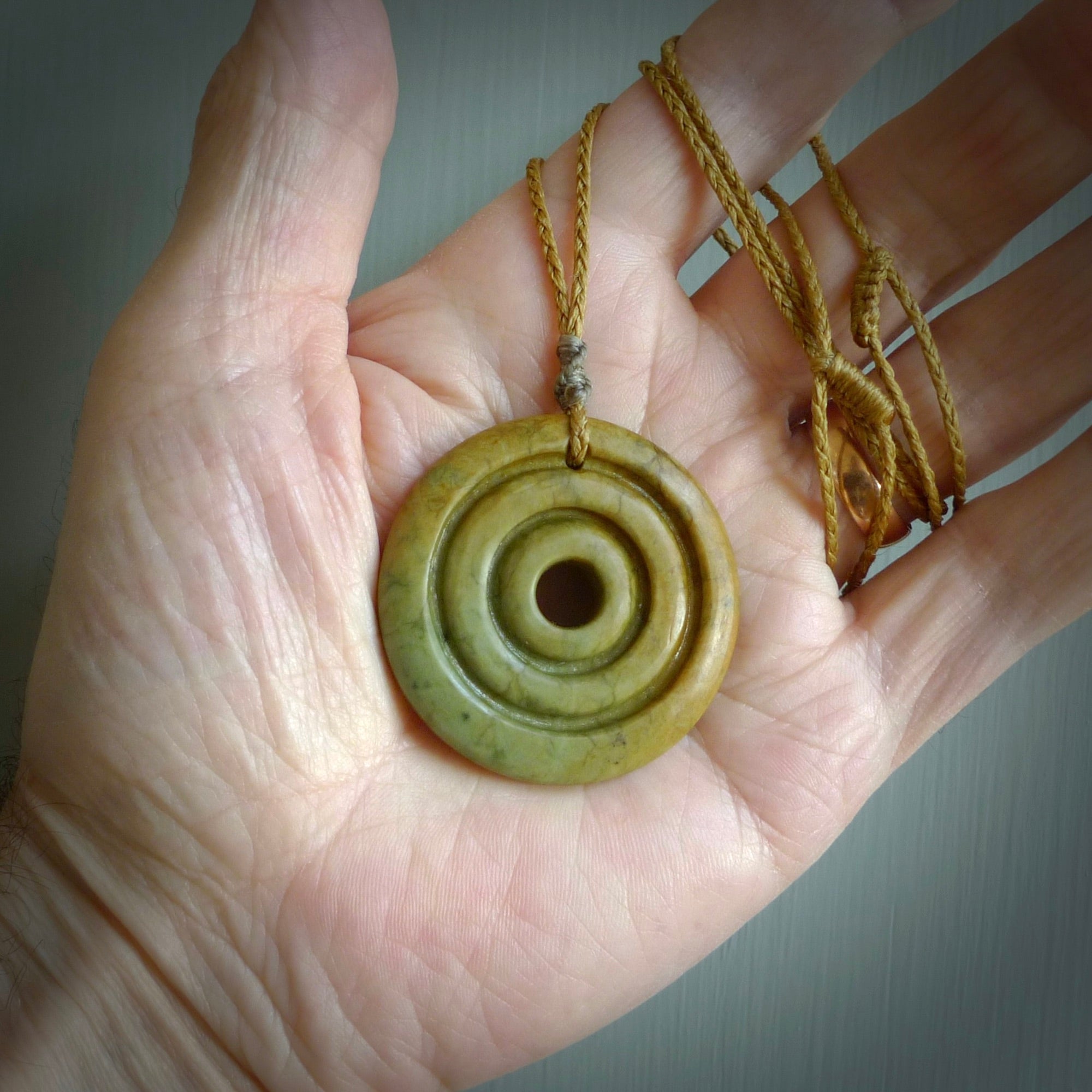 This picture shows a hand carved disc pendant made from a bright orange and green coloured flower jade. The front face is a unique design and the back is flat. It is provided with an adjustable kowhai yellow cord.