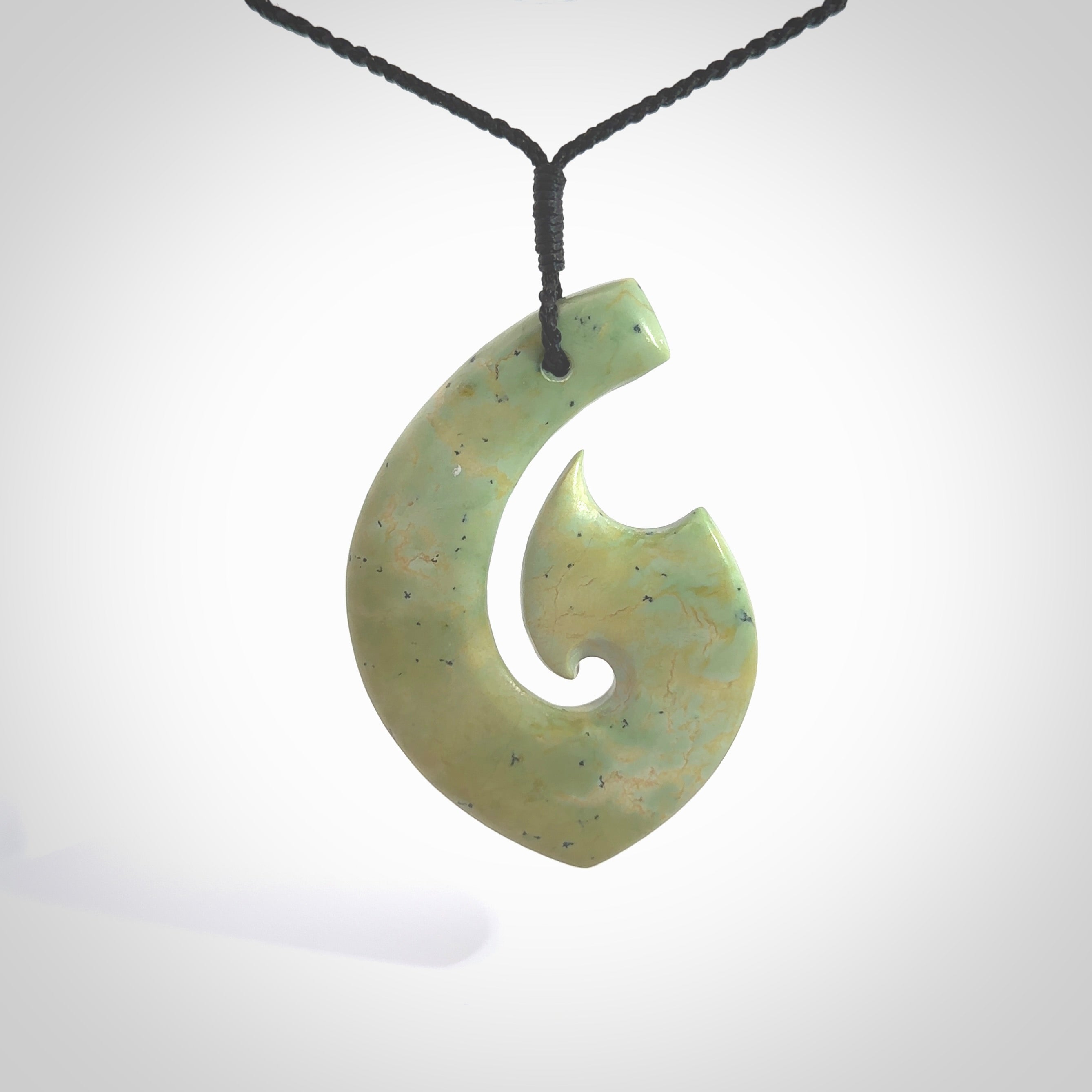 This picture shows a carved Matau, Hook, in New Zealand Jade. It is a hook shaped pendant pendant that culminates in a fish tail. It is provided with a black coloured cord that is length adjustable. Hand made in New Zealand by Andrew Doughty.