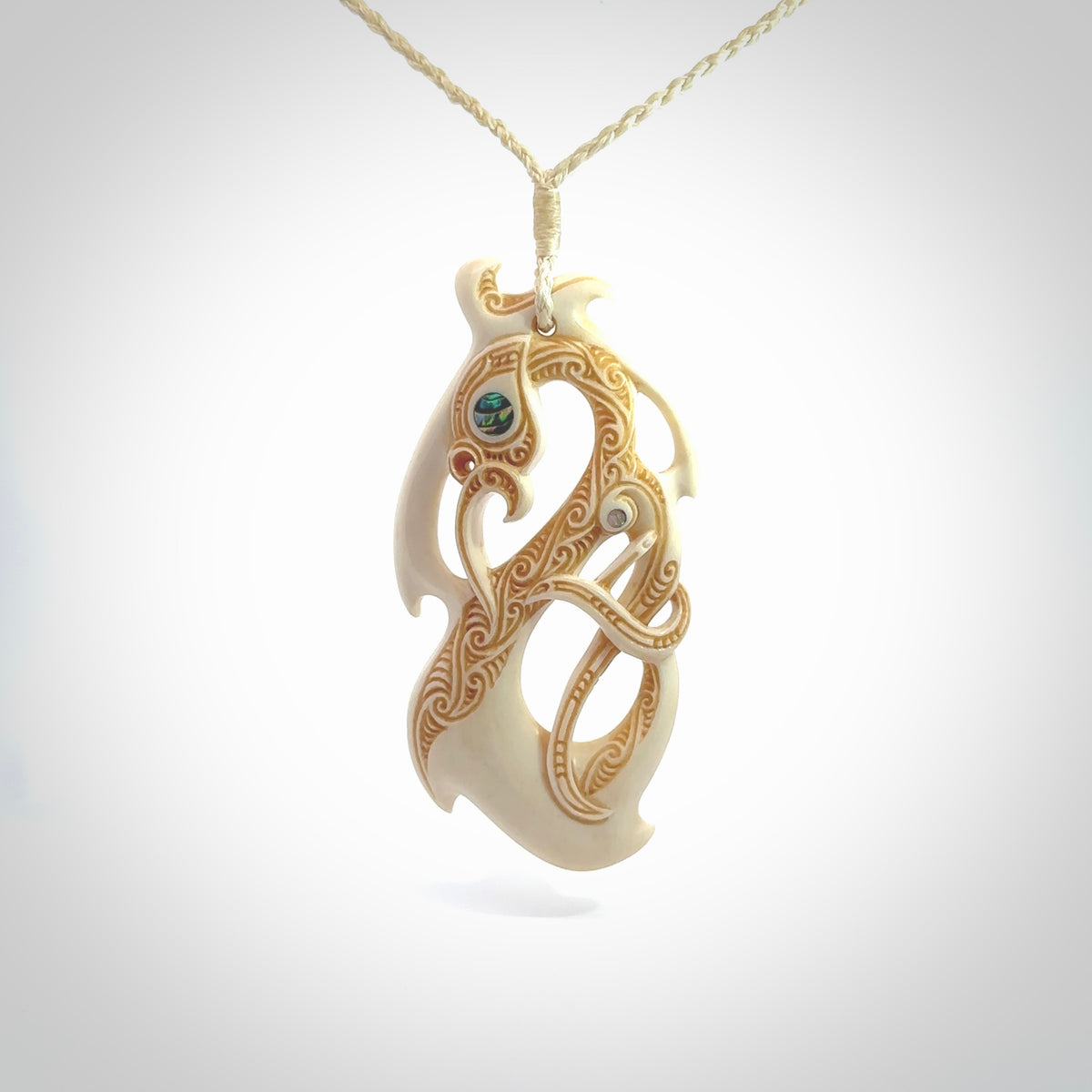 This picture shows a carved manaia in bone. It is a manaia shaped pendant that has great detail. The artist has carved traditional decorative Koru designs into the side of the body and these run up the sides of the manaia head. These have specific meanings. It is provided with a hand-plaited beige cord that is length adjustable.