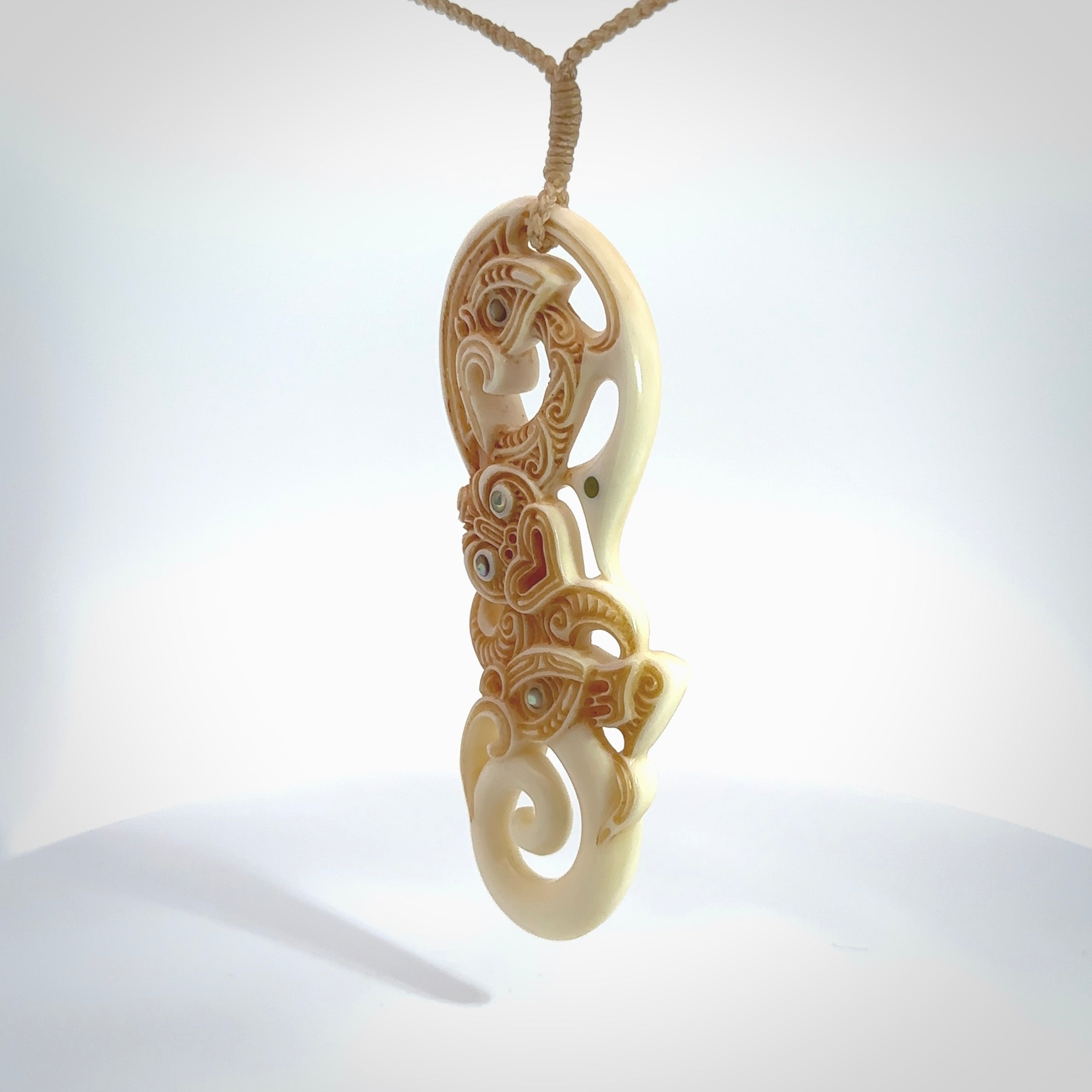 This picture shows a carved manaia in bone. It has a Tiki face on the front that culminates in a koru. The artist has carved traditional decorative Koru designs into the side of the body and these run up the sides of the manaia head. These have specific meanings. It is provided with a hand-plaited beige cord that is length adjustable.