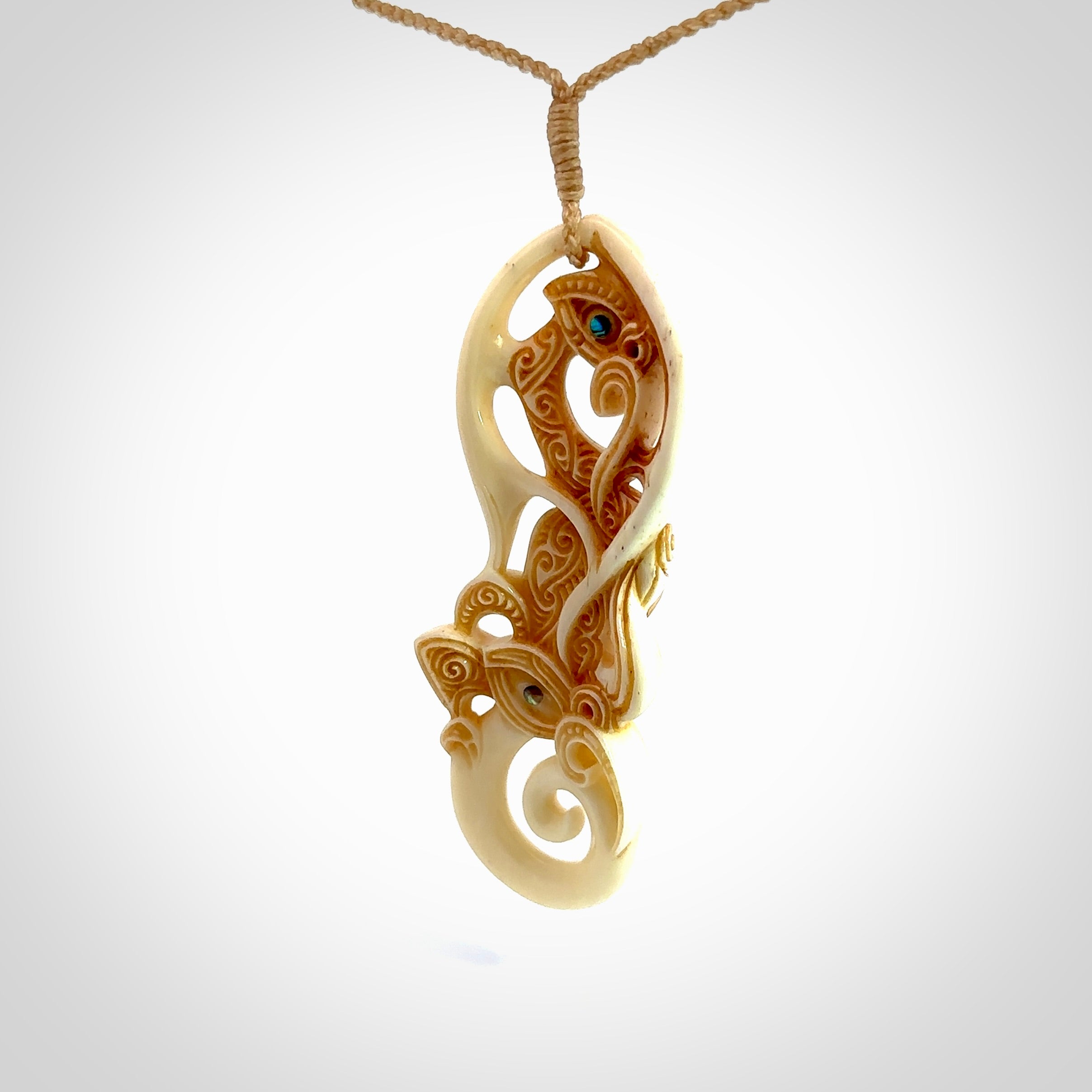 This picture shows a carved manaia in bone. It has a Tiki face on the front that culminates in a koru. The artist has carved traditional decorative Koru designs into the side of the body and these run up the sides of the manaia head. These have specific meanings. It is provided with a hand-plaited beige cord that is length adjustable.