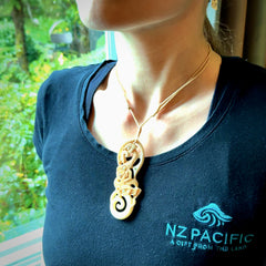 This picture shows a carved manaia in bone. It has a Tiki face on the front that culminates in a koru. The artist has carved traditional decorative Koru designs into the side of the body and these run up the sides of the manaia head. These have specific meanings. It is provided with a hand-plaited beige cord that is length adjustable.