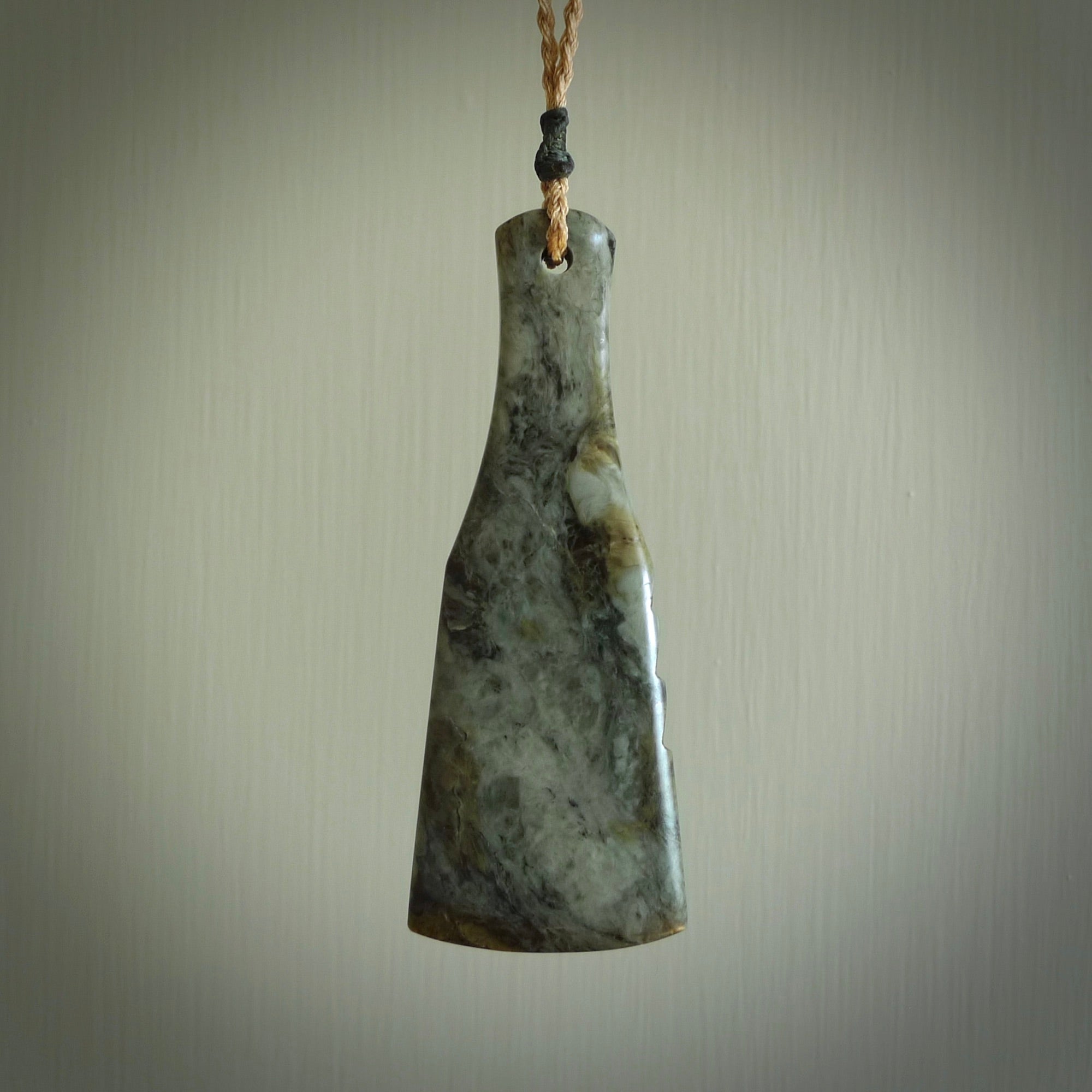 This is a sleek and slender hand carved New Zealand Jade mere pendant. The stone is a very dark green with mottled pale inclusions. The cord is tan and adjustable. The pendant has a light polish and just glows.