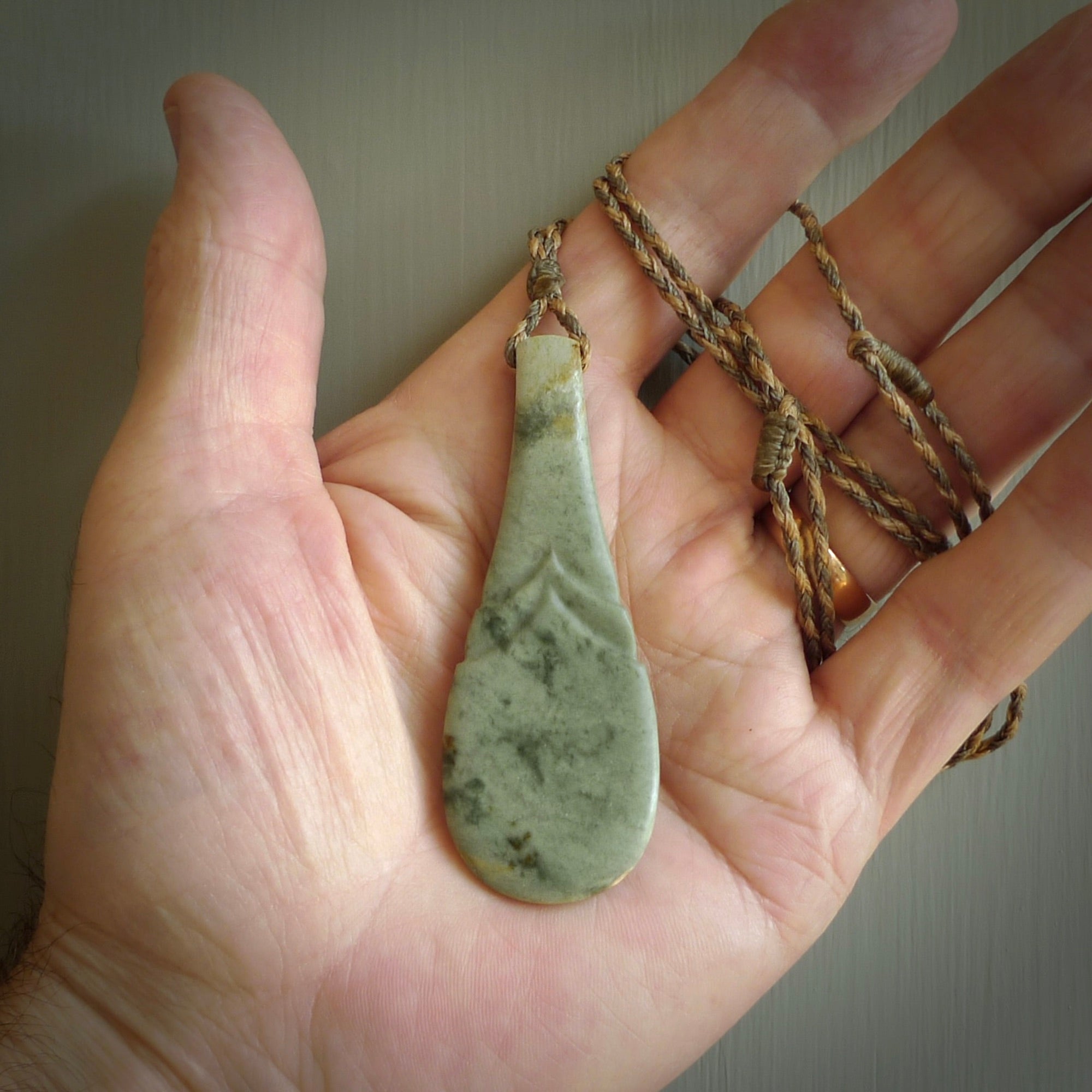 This is a sleek and slender hand carved New Zealand Jade mere pendant. The stone is a very pale green with mottled pale inclusions. The cord is tan and khaki and adjustable. The pendant is delivered in a woven kete pouch.