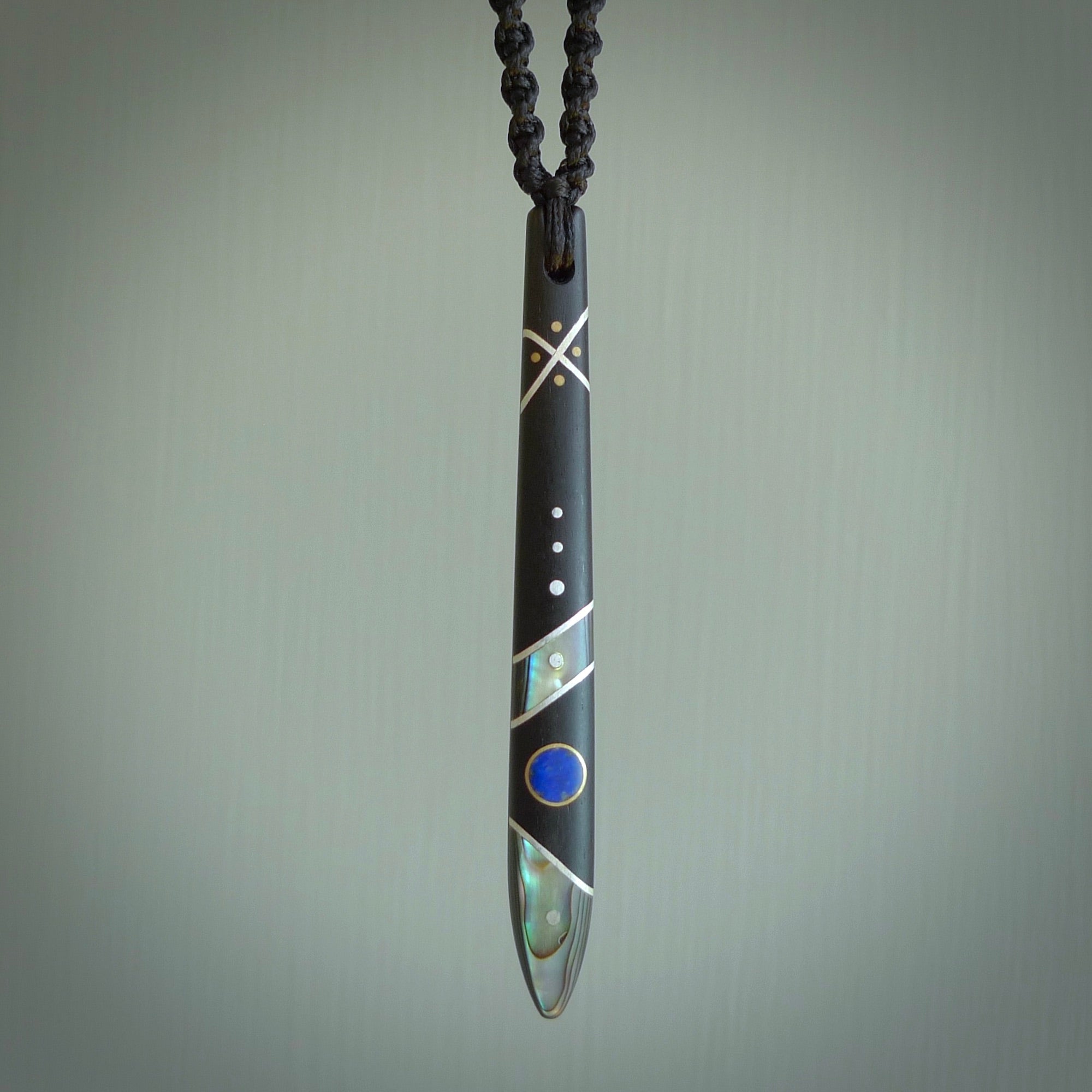 This photo shows a medium sized taiaha pendant hand carved from piano key with Afghani Lapis Lazuli and Chinese Glowing Stone, Sleeping Beauty Turquoise inlay alongside; brass and silver. This is a stand out one off necklace for those who appreciate art to wear. It is provided with a cord in black that is a fixed length with Paua Shell Toggle. We ship this piece worldwide and shipping is included in the price.