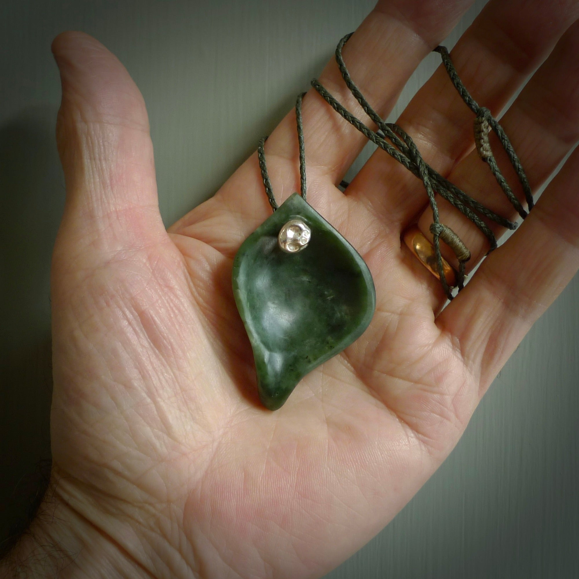 This picture shows a hand carved drop pendant made from a deep green coloured New Zealand Pounamu, jade. The front face is a concave, cupped design and the back is round. It is provided with an adjustable fern green cord in a woven kete pouch. Shipped to you with Express Courier.