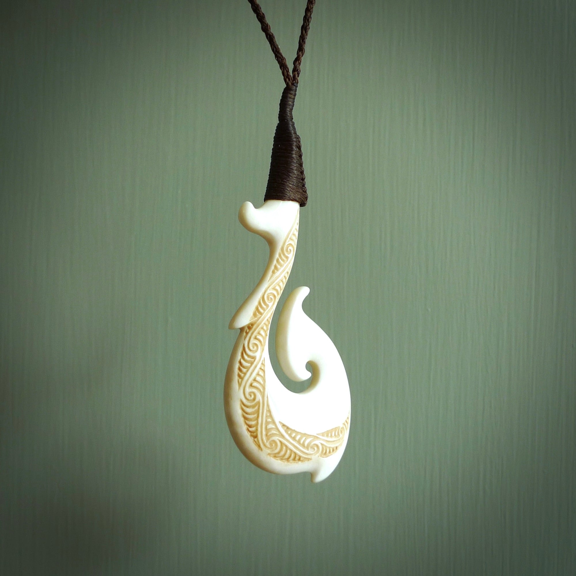 This picture shows a carved hook in bone. The artist has carved traditional decorative kowhaiwhai designs into the body and these run up the insides of the matau. These have specific meanings. It is provided with a hand-plaited brown cord that is length adjustable. Free shipping worldwide.