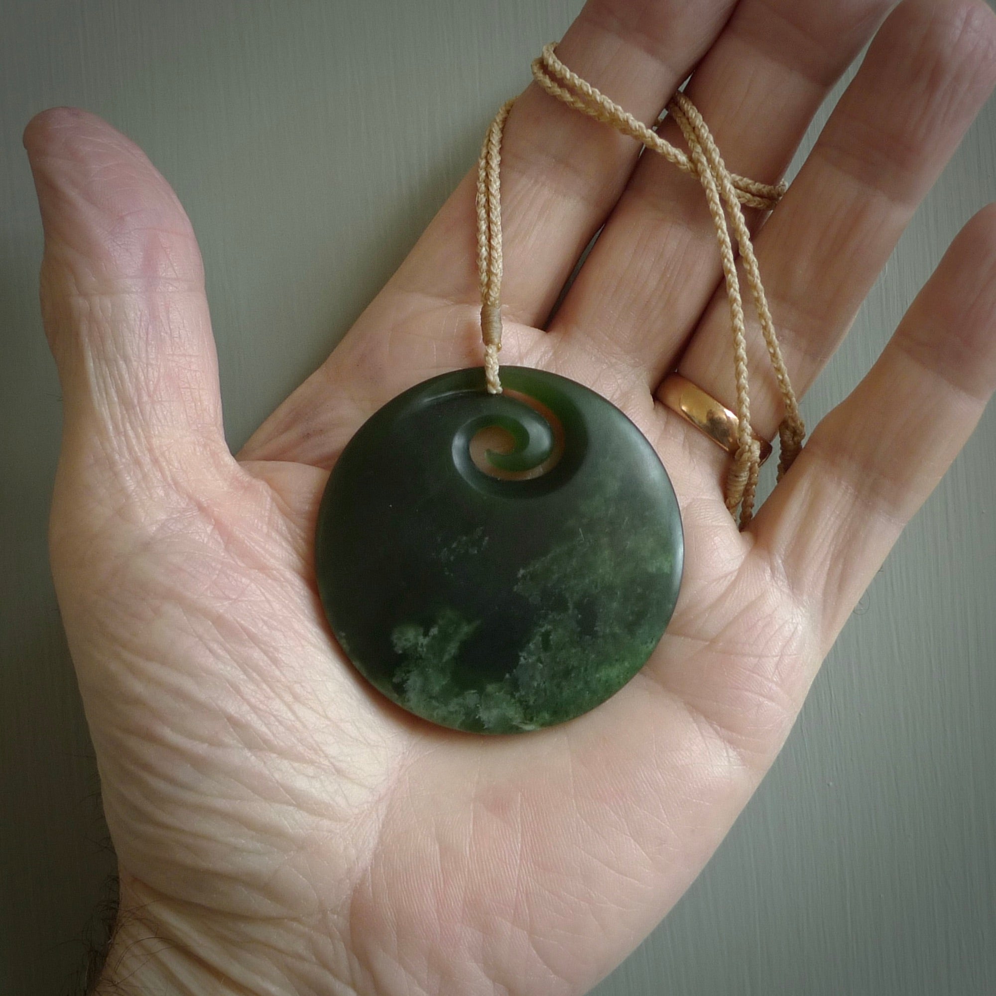 This picture shows a large hand carved jade koru pendant. It is a darker green colour which is quite translucent where the jade is thin. Although a large piece, it is a wonderful, delicate piece of jewellery. The cord is hand plaited and adjustable so that you can position the pendant where it suits you best. This koru was carved for us by Ric Moor. Delivery is free worldwide.