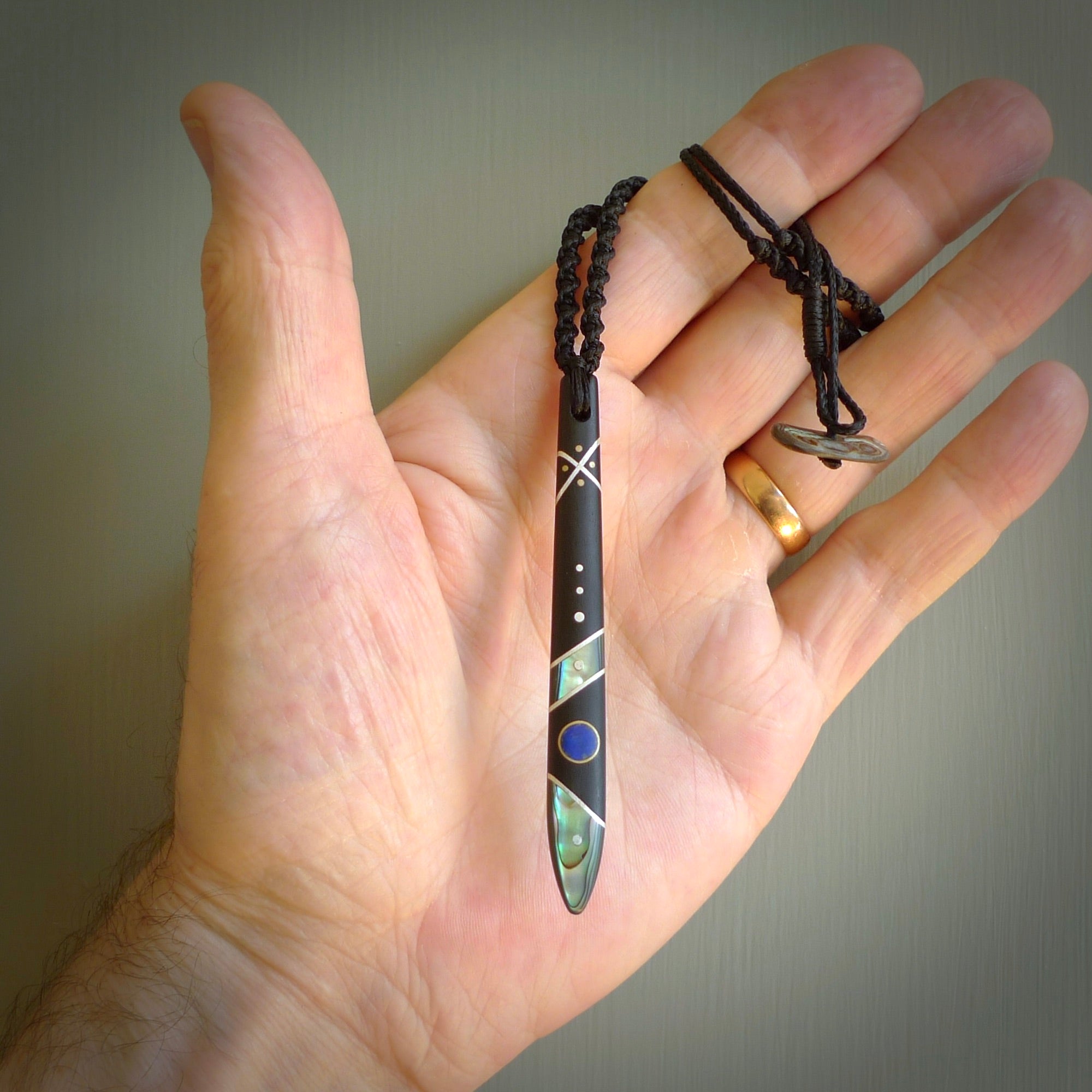 This photo shows a medium sized taiaha pendant hand carved from piano key with Afghani Lapis Lazuli and Chinese Glowing Stone, Sleeping Beauty Turquoise inlay alongside; brass and silver. This is a stand out one off necklace for those who appreciate art to wear. It is provided with a cord in black that is a fixed length with Paua Shell Toggle. We ship this piece worldwide and shipping is included in the price.