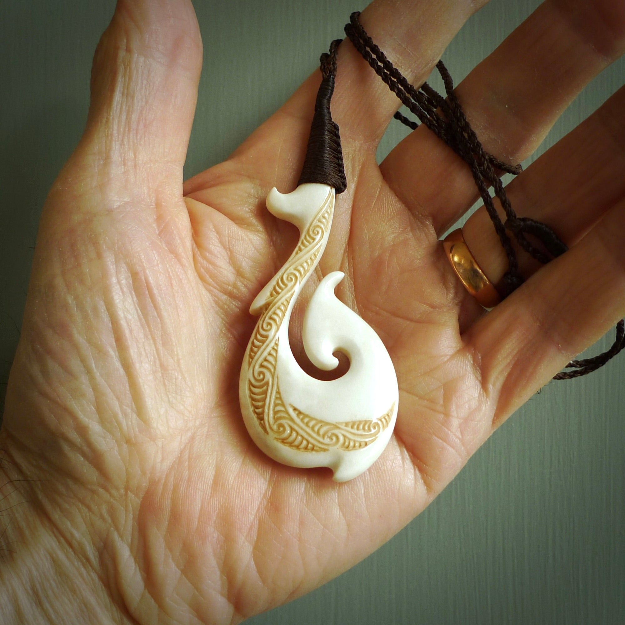 This picture shows a carved hook in bone. The artist has carved traditional decorative kowhaiwhai designs into the body and these run up the insides of the matau. These have specific meanings. It is provided with a hand-plaited brown cord that is length adjustable. Free shipping worldwide.