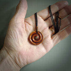 A hand carved and intricate koru pendant made for us by Yuri Terenyi. This is a beautiful little piece and is emblematic of the well known and loved Koru design. It is carved from stained bone in a hollowed, koru spiral shape with paua shell inlay in the middle of the koru. It is suspended from a black cord with a gingernut floret and the necklace is adjustable.