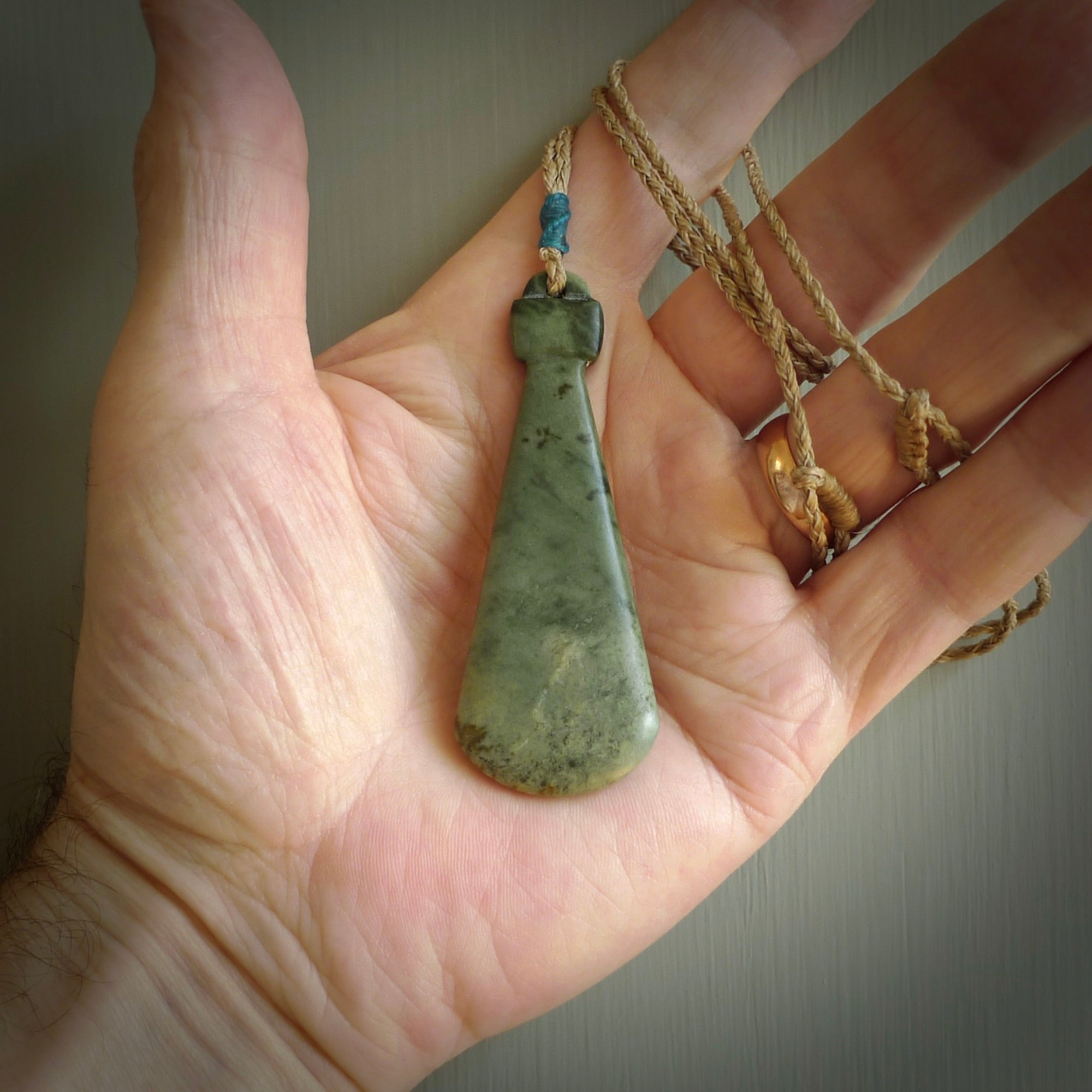 This is a sleek and slender hand carved New Zealand Jade mere pendant. The stone is a light green with mottled inclusions. The cord is tan and adjustable. The pendant has a light polish and just glows. Delivered in a woven kete pouch.