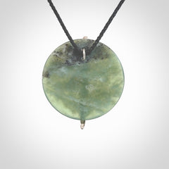 Hand carved New Zealand Inanga circle disc drop with sterling silver shard pendant. Made by Ana Krakosky for NZ Pacific in New Zealand Pounamu, jade. A beautiful light and varied green Inanga Pounamu, jade medium sized circle disc, drop necklace with silver cast insert, provided on a black adjustable cord.