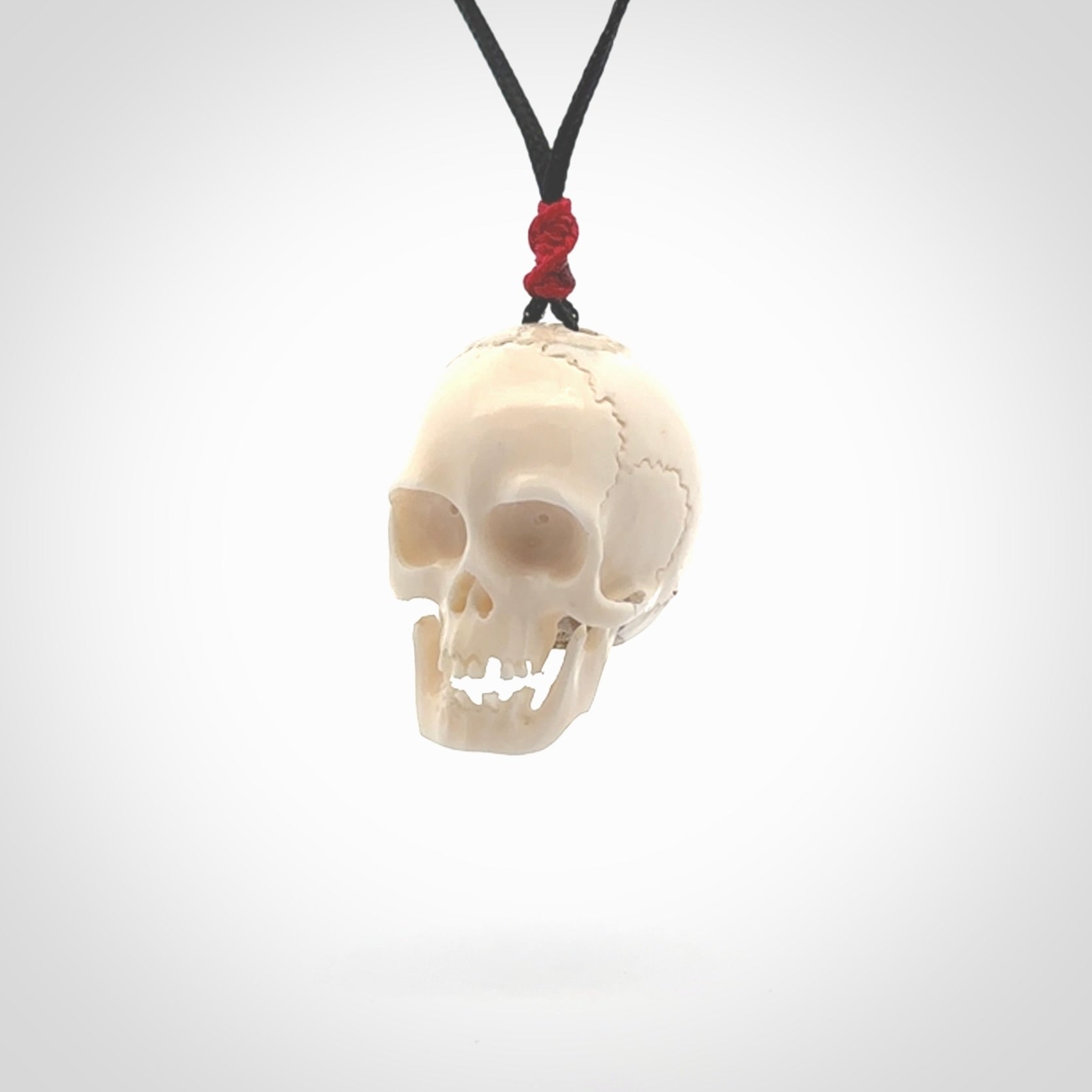 This picture shows a large sized Deer Antler Bone skull pendant that we have hand carved. It is polished to a soft shine and is a very striking piece. The cord is black and has a small red popper binding just above the pendant.