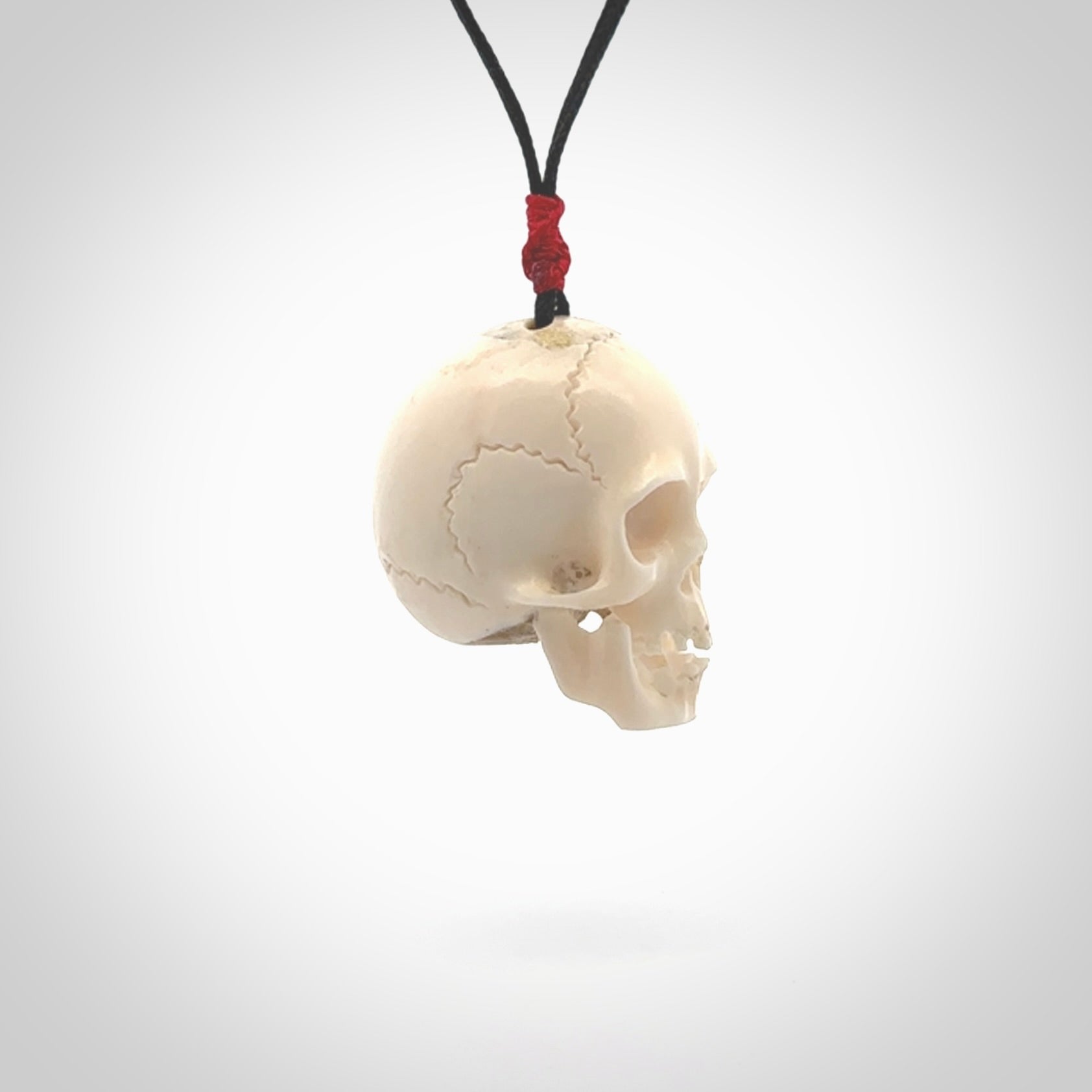 This picture shows a large sized Deer Antler Bone skull pendant that we have hand carved. It is polished to a soft shine and is a very striking piece. The cord is black and has a small red popper binding just above the pendant.