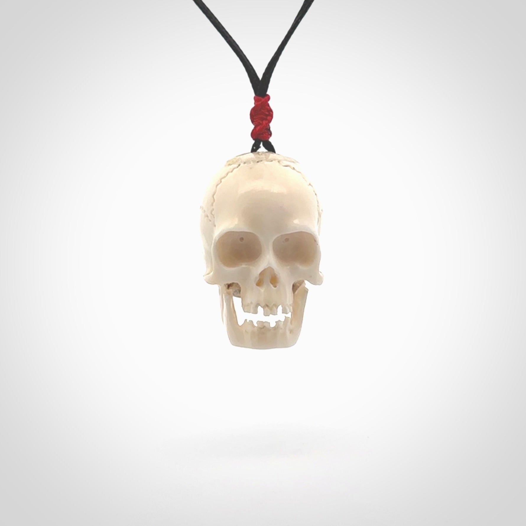 This picture shows a large sized Deer Antler Bone skull pendant that we have hand carved. It is polished to a soft shine and is a very striking piece. The cord is black and has a small red popper binding just above the pendant.