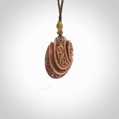 A hand carved and intricate koru pendant made for us by Yuri Terenyi. This is a beautiful little piece and is emblematic of the well known and loved Koru design. It is carved from bone in a hollowed, oval ball shape with decorative design carved into the koru. It is suspended from a chocolate brown cord which is adjustable.