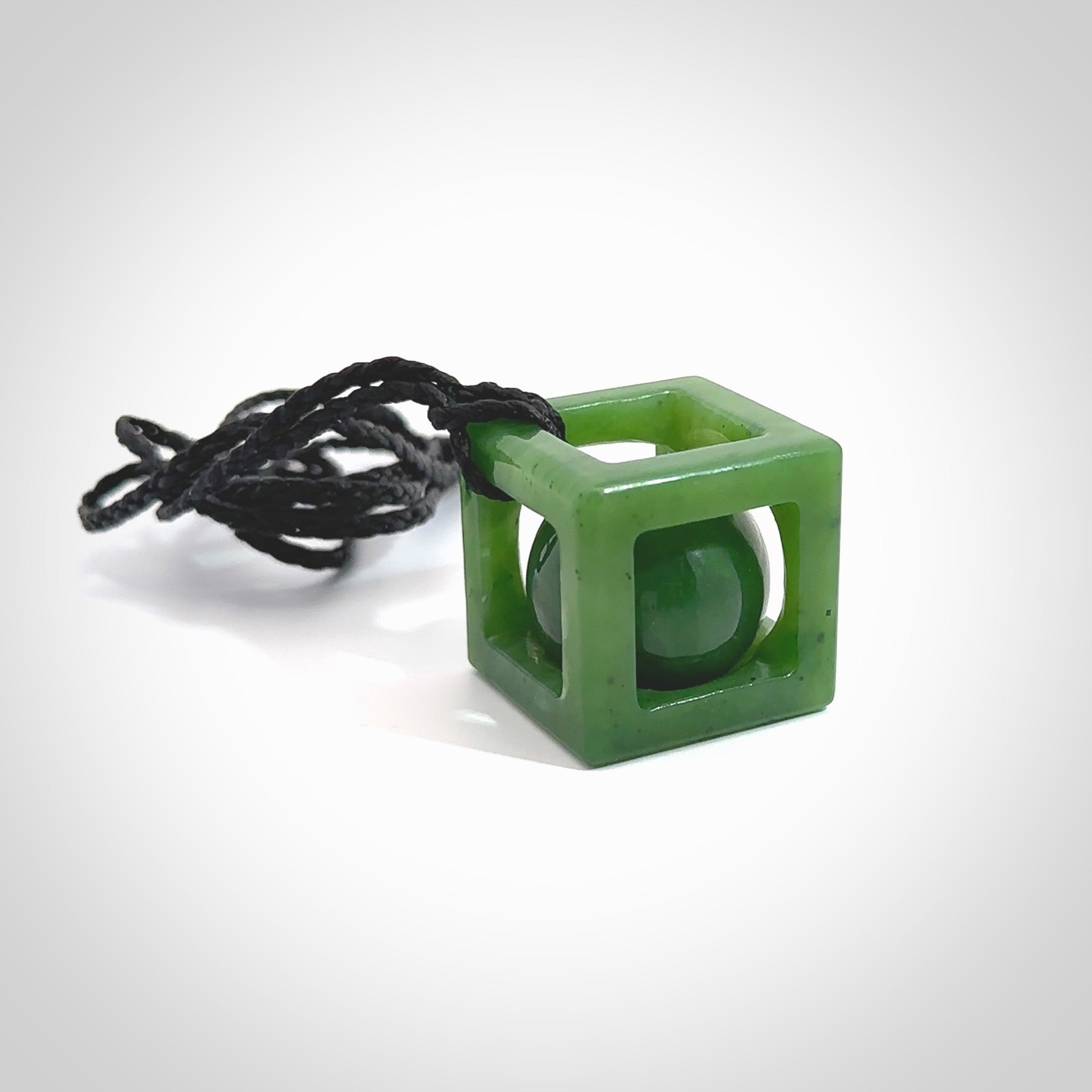 A hand carved medium sized green Jade square box with ball inside necklace. The cord is black and adjustable in length. A medium sized hand made cube/box with ball necklace in green Jade. One off work of art to wear.