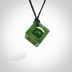 A hand carved medium sized green Jade square box with ball inside necklace. The cord is black and adjustable in length. A medium sized hand made cube/box with ball necklace in green Jade. One off work of art to wear.