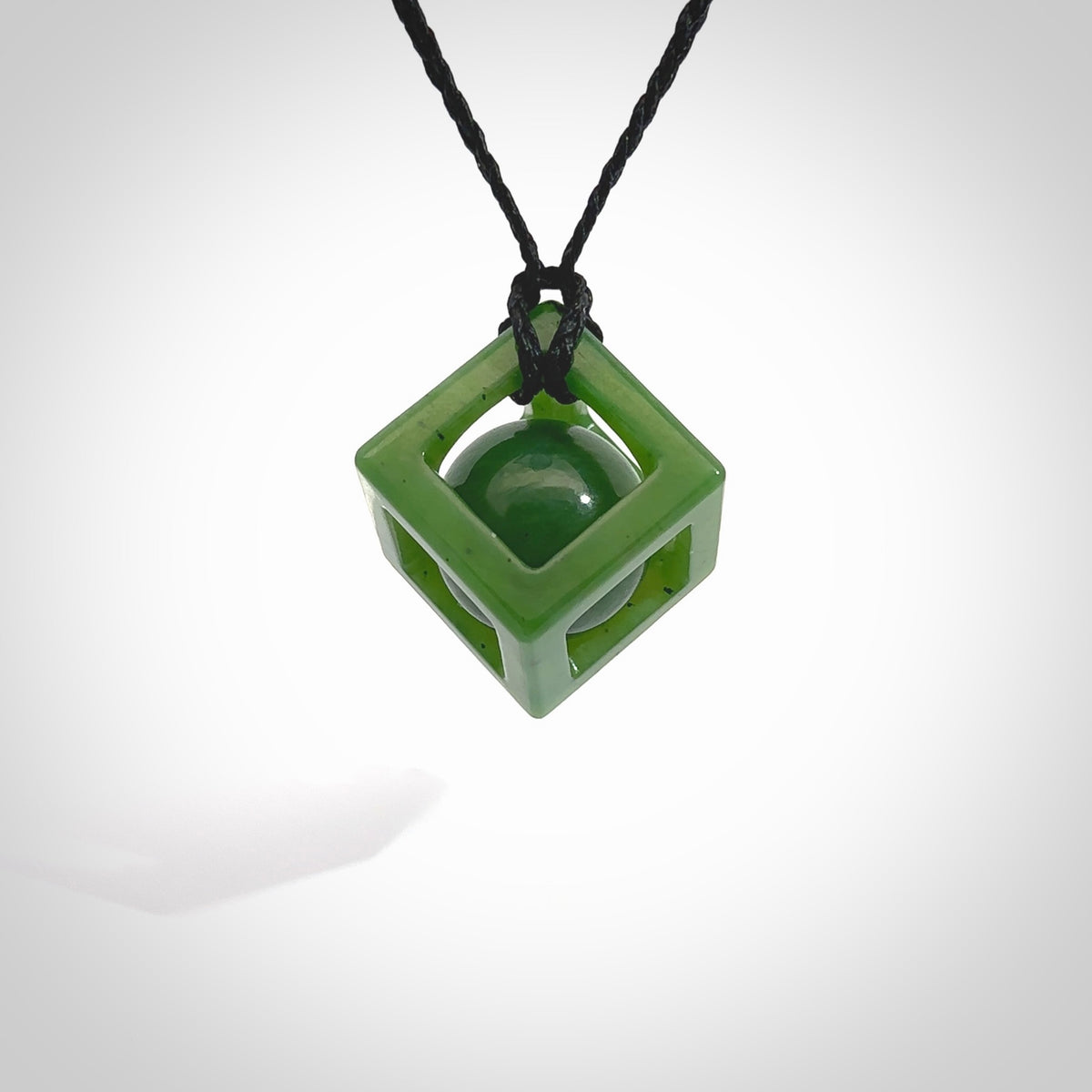 A hand carved medium sized green Jade square box with ball inside necklace. The cord is black and adjustable in length. A medium sized hand made cube/box with ball necklace in green Jade. One off work of art to wear.