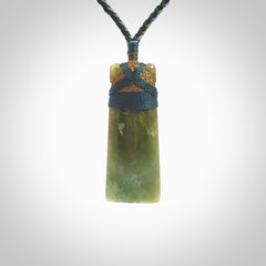 This photo shows a beautiful New Zealand jade toki suspended from a hand plaited adjustable cord. The binding around the top of the toki is a blue and brown colour. The cord is length adjustable which allows the pendant to be positioned where it suits you best. The toki was carved for us by Jen Hung who is a wonderful jade carver from New Zealand.