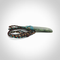 This photo shows a beautiful New Zealand jade toki suspended from a hand plaited adjustable cord. The binding around the top of the toki is a blue and bronze colour. The cord is length adjustable which allows the pendant to be positioned where it suits you best. The toki was carved for us by Jen Hung who is a wonderful jade carver from New Zealand.