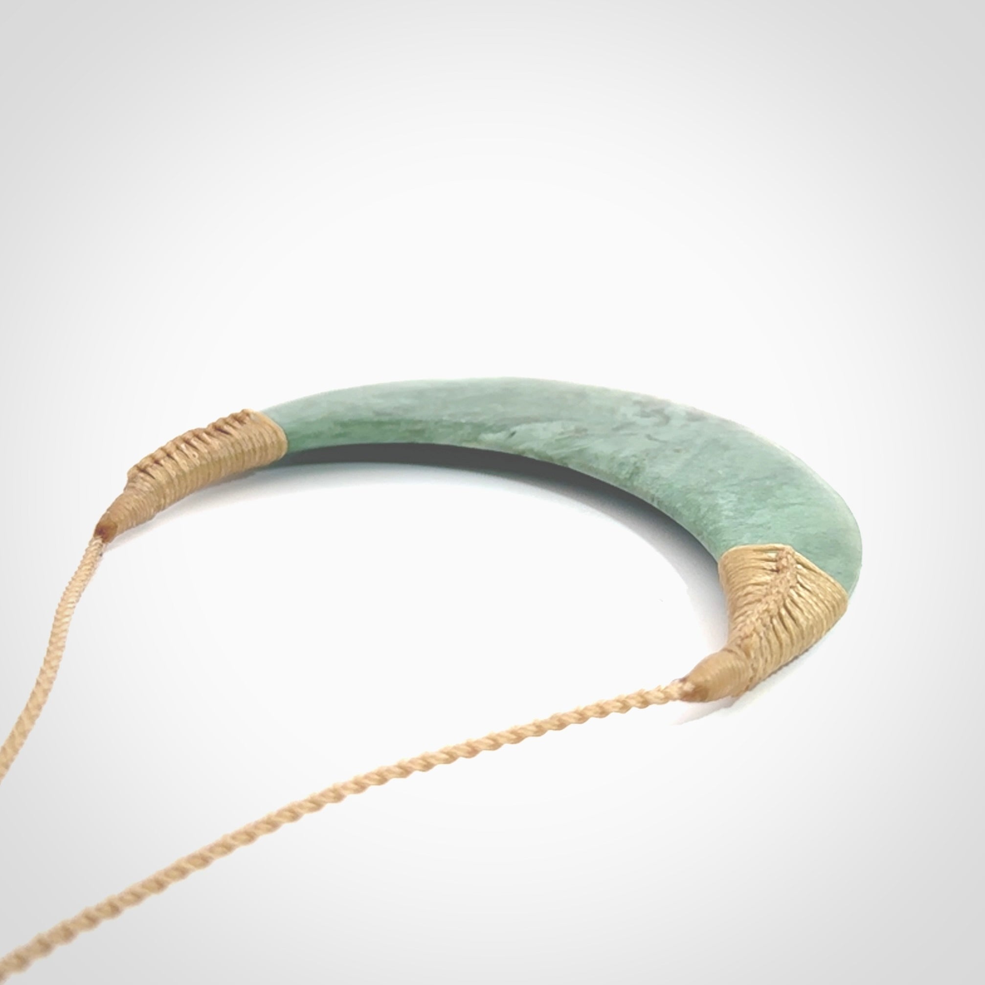 This picture shows a hand carved New Zealand Inanga jade contemporary half-shield pendant. It is a light blue and green colour and is a wonderful, large piece of jewellery. The cord is hand plaited and adjustable so that you can position the pendant where it suits you best. This piece was carved for us by Kyohei Noguchi. Delivery is free worldwide.