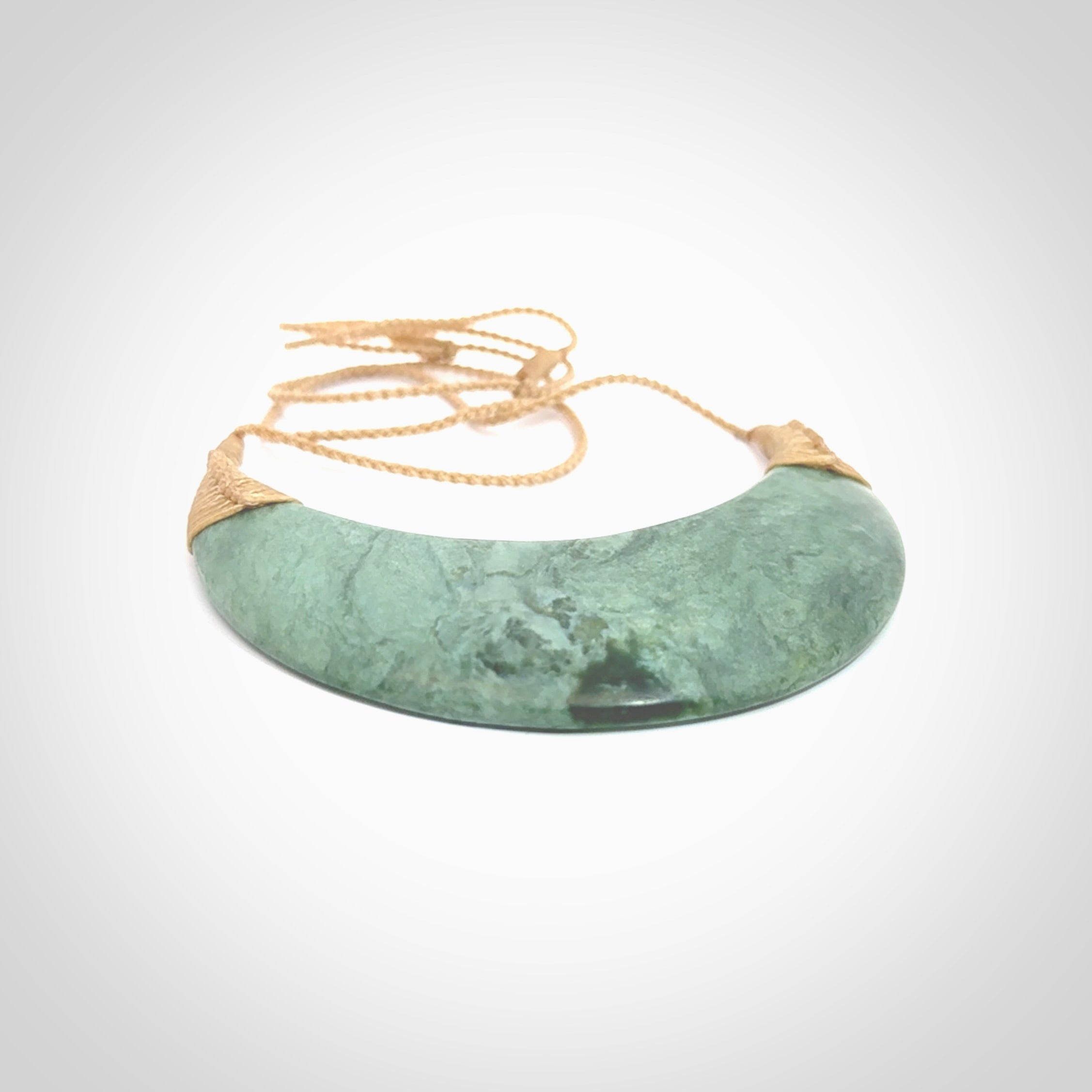 This picture shows a hand carved New Zealand Inanga jade contemporary half-shield pendant. It is a light blue and green colour and is a wonderful, large piece of jewellery. The cord is hand plaited and adjustable so that you can position the pendant where it suits you best. This piece was carved for us by Kyohei Noguchi. Delivery is free worldwide.