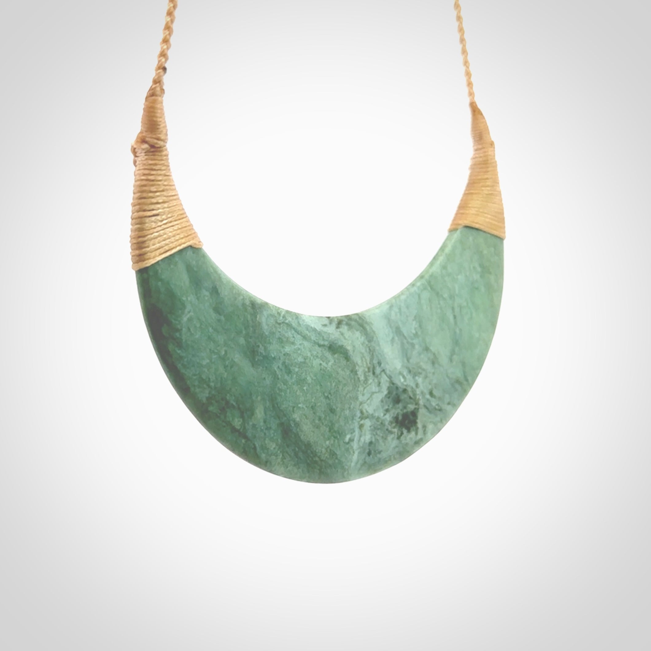 This picture shows a hand carved New Zealand Inanga jade contemporary half-shield pendant. It is a light blue and green colour and is a wonderful, large piece of jewellery. The cord is hand plaited and adjustable so that you can position the pendant where it suits you best. This piece was carved for us by Kyohei Noguchi. Delivery is free worldwide.